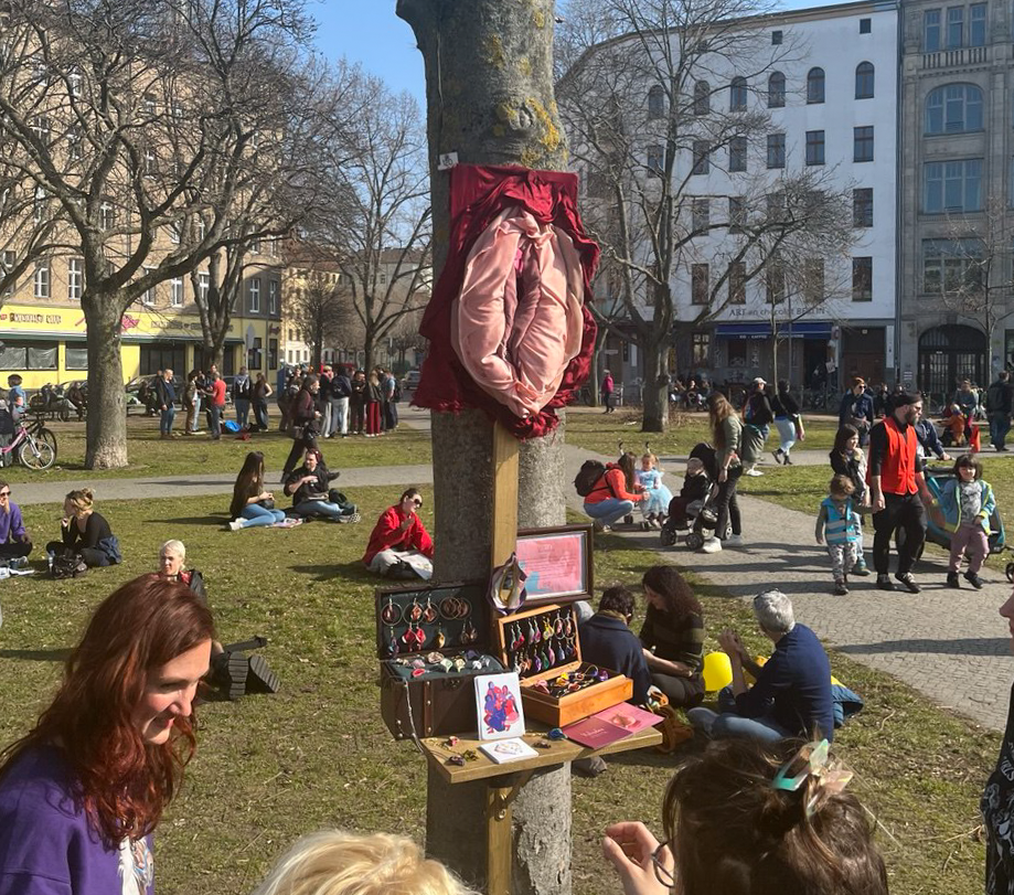 A bazaar or market setup with jewelry displayed on a stand attached to a tree in an outdoor park. There are numerous people sitting and walking in the background, with some trees and buildings visible, and clear weather.
