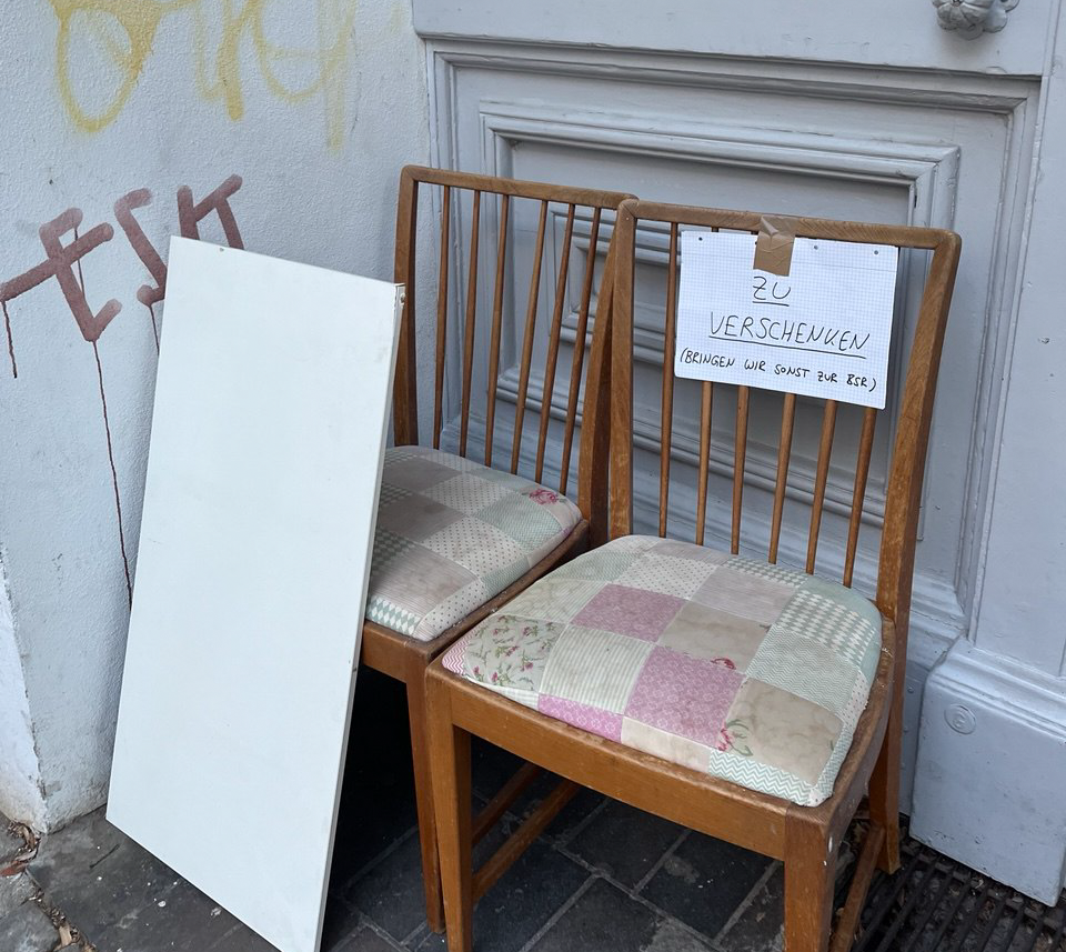 Two wooden chairs with patchwork cushions and a white sign taped to one chair, placed against a white wall with graffiti.