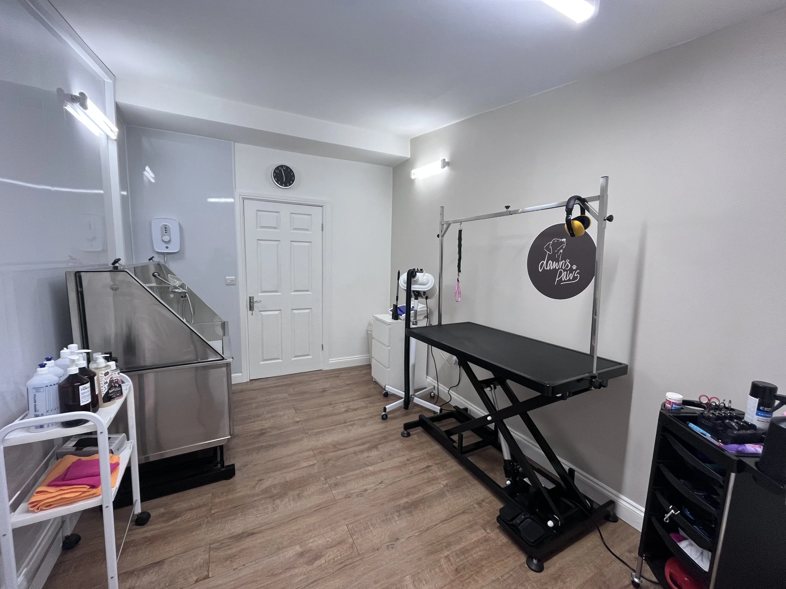 A veterinary clinic or grooming room with a grooming table, grooming tools, and supplies, with white walls and wooden flooring.