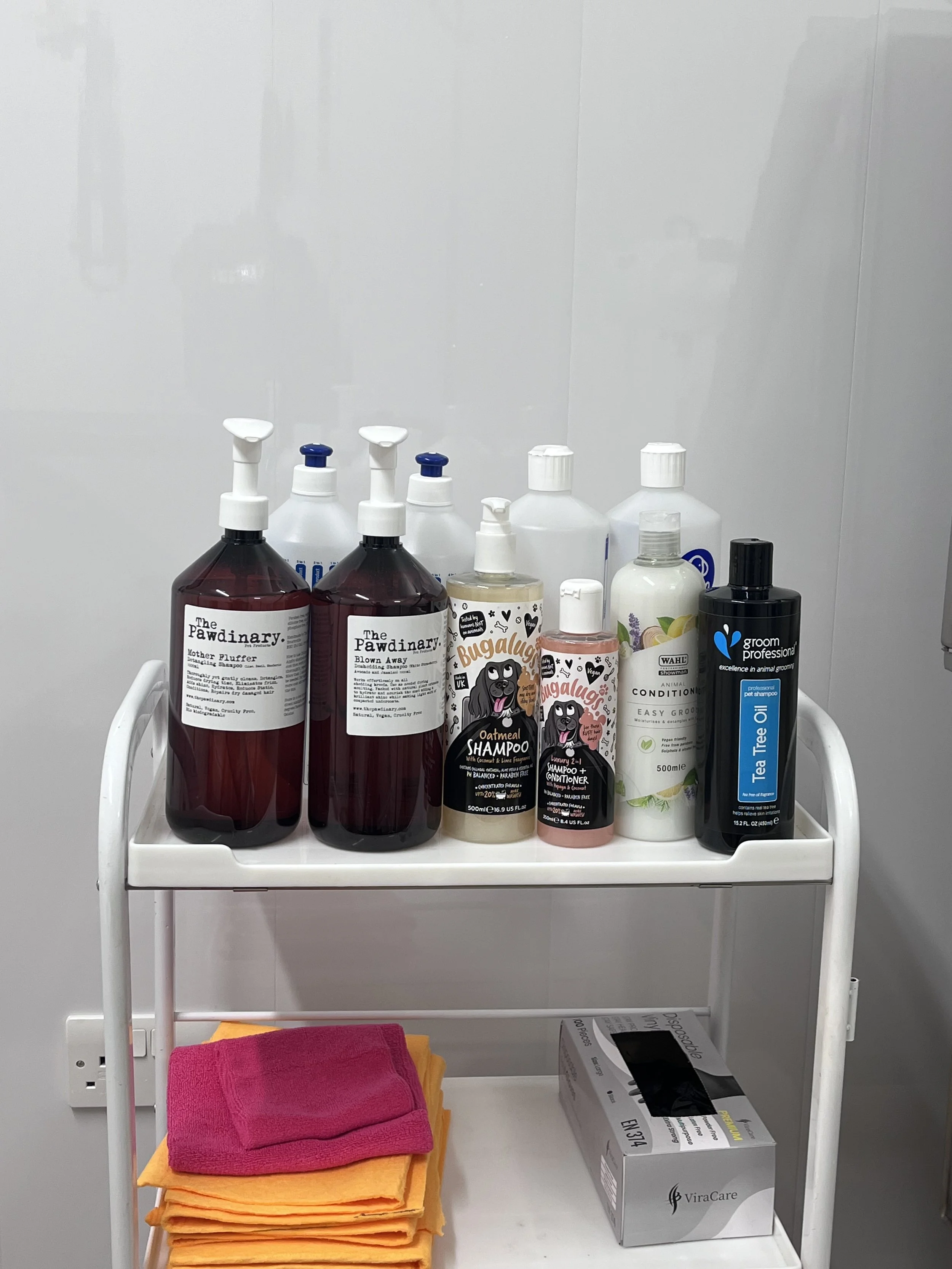 A white shelves with bottles of dog grooming supplies, including shampoos, conditioners, and tea tree oil, along with folded towels and a box of disposable gloves.