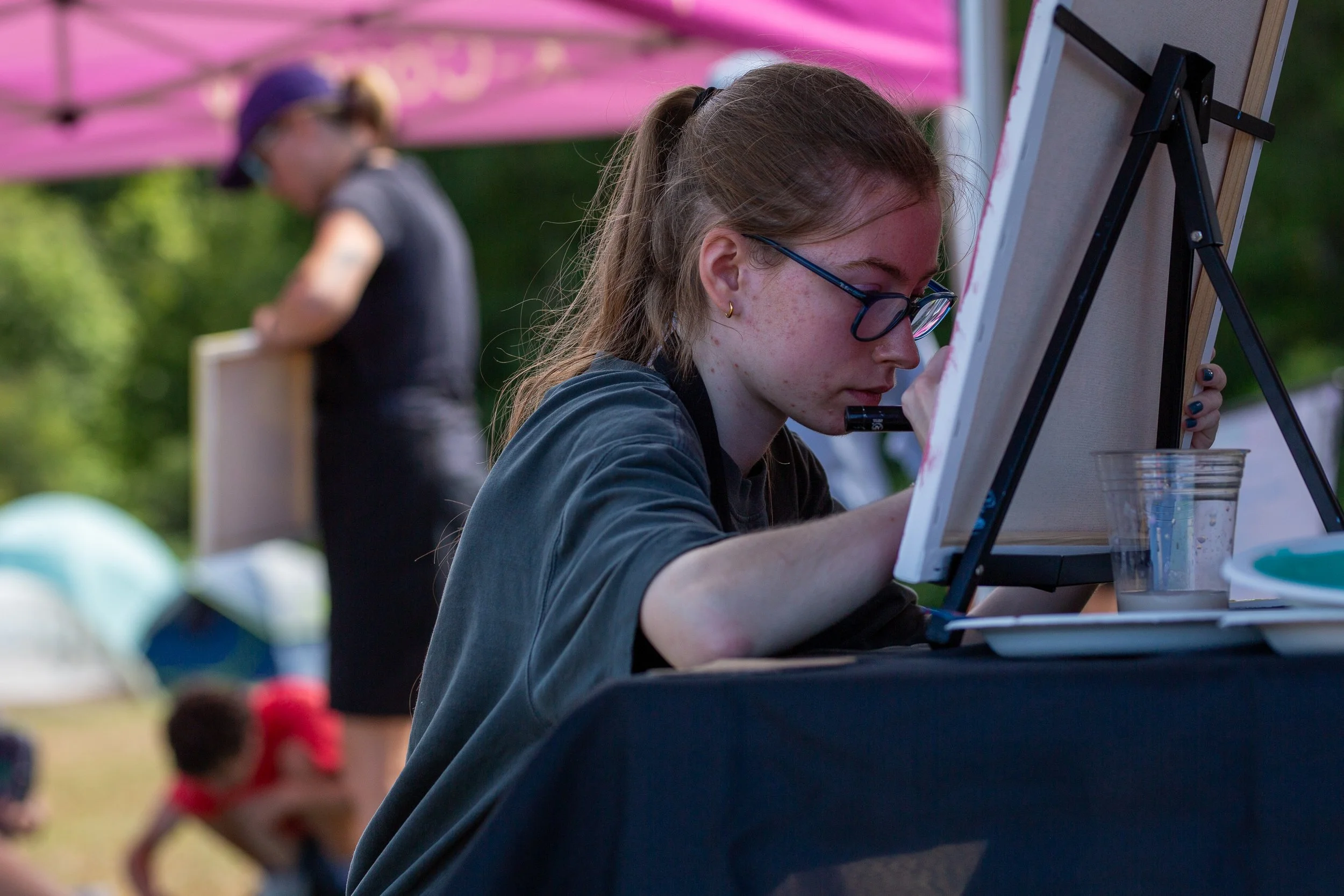 A teenage girl with glasses and freckles painting on a canvas outdoors with an easel, while another person in the background appears to be painting, under a pink umbrella.