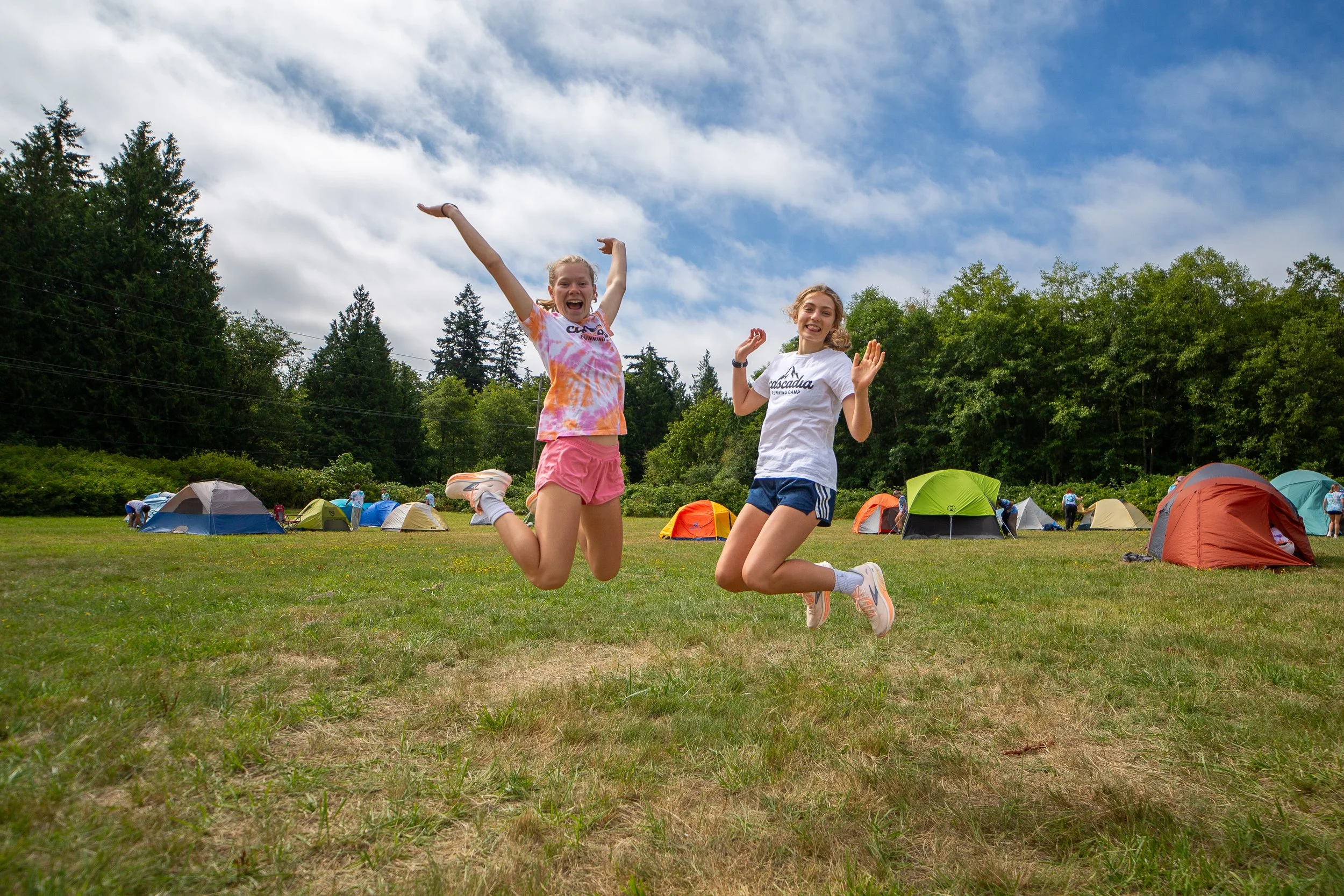 Two girls are jumping in the air at a campground with colorful tents, green trees, and a partly cloudy sky in the background.
