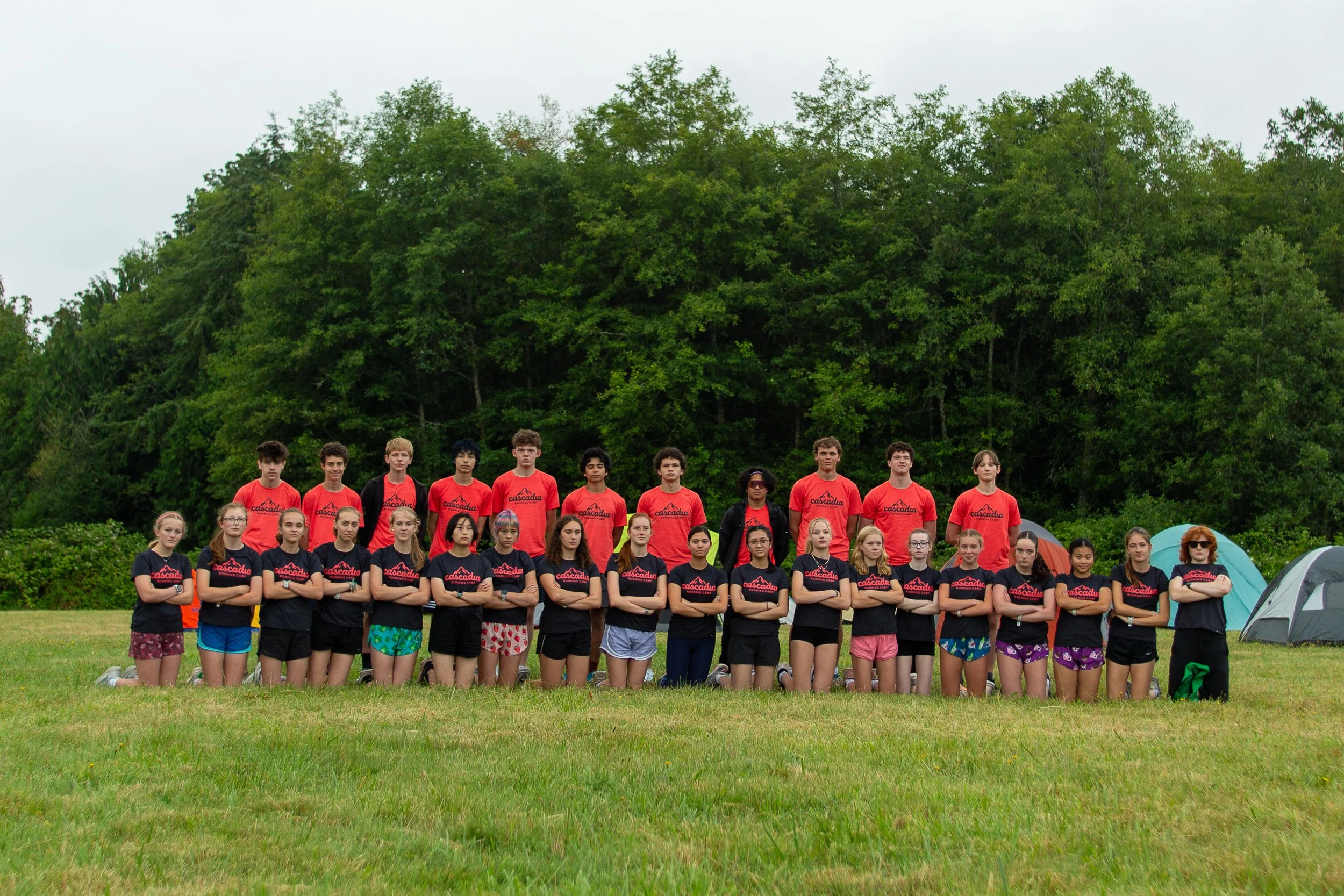 Group of children and teenagers standing outdoors on grass, with a backdrop of trees and tents, some wearing matching dark shirts with red and white logos and others in colorful shorts and tshirts.