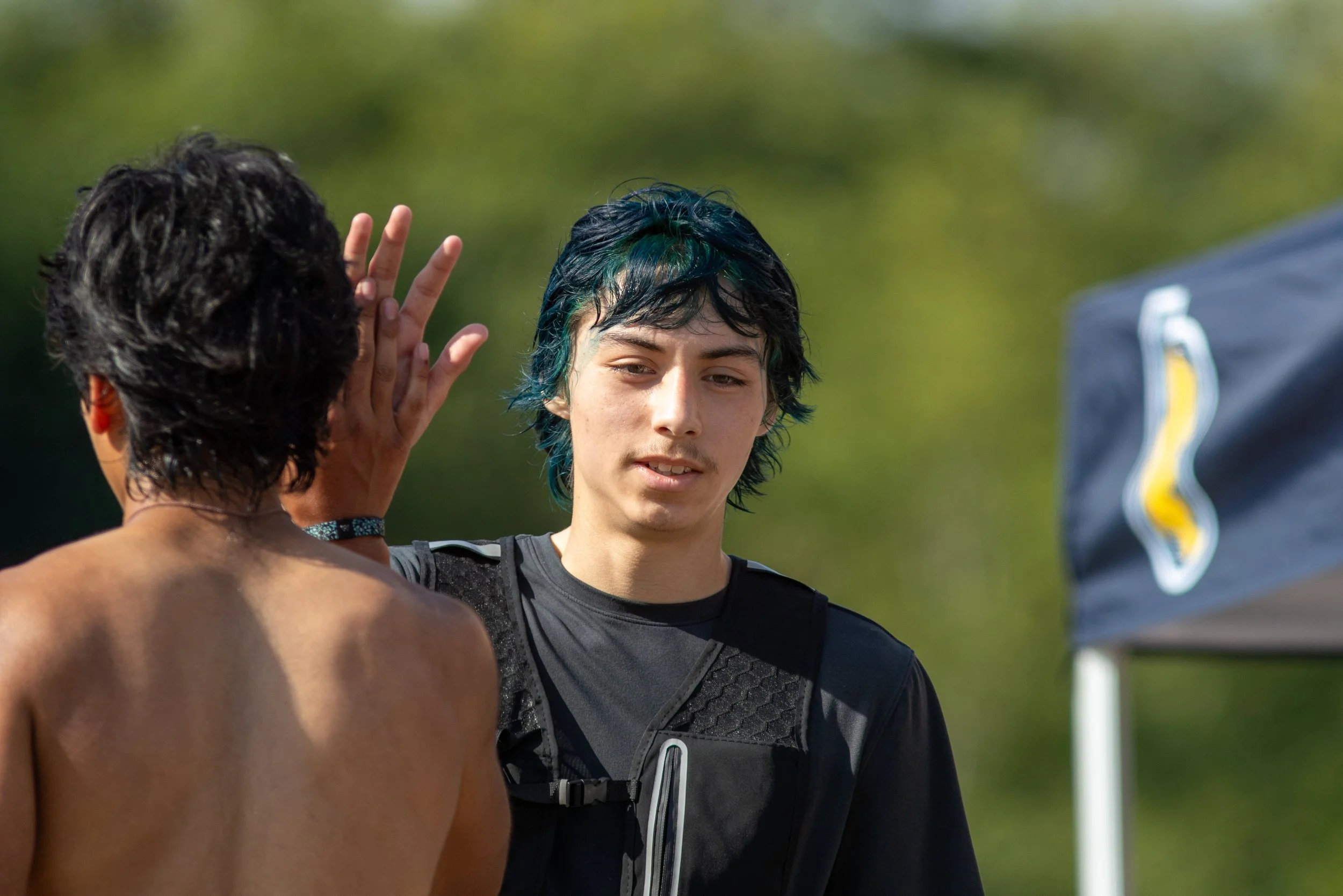 Two young men, one with dark curly hair and the other with short black hair streaked with blue, seemingly engaged in a conversation outdoors on a sunny day, with a blurred green background and an American football nearby.