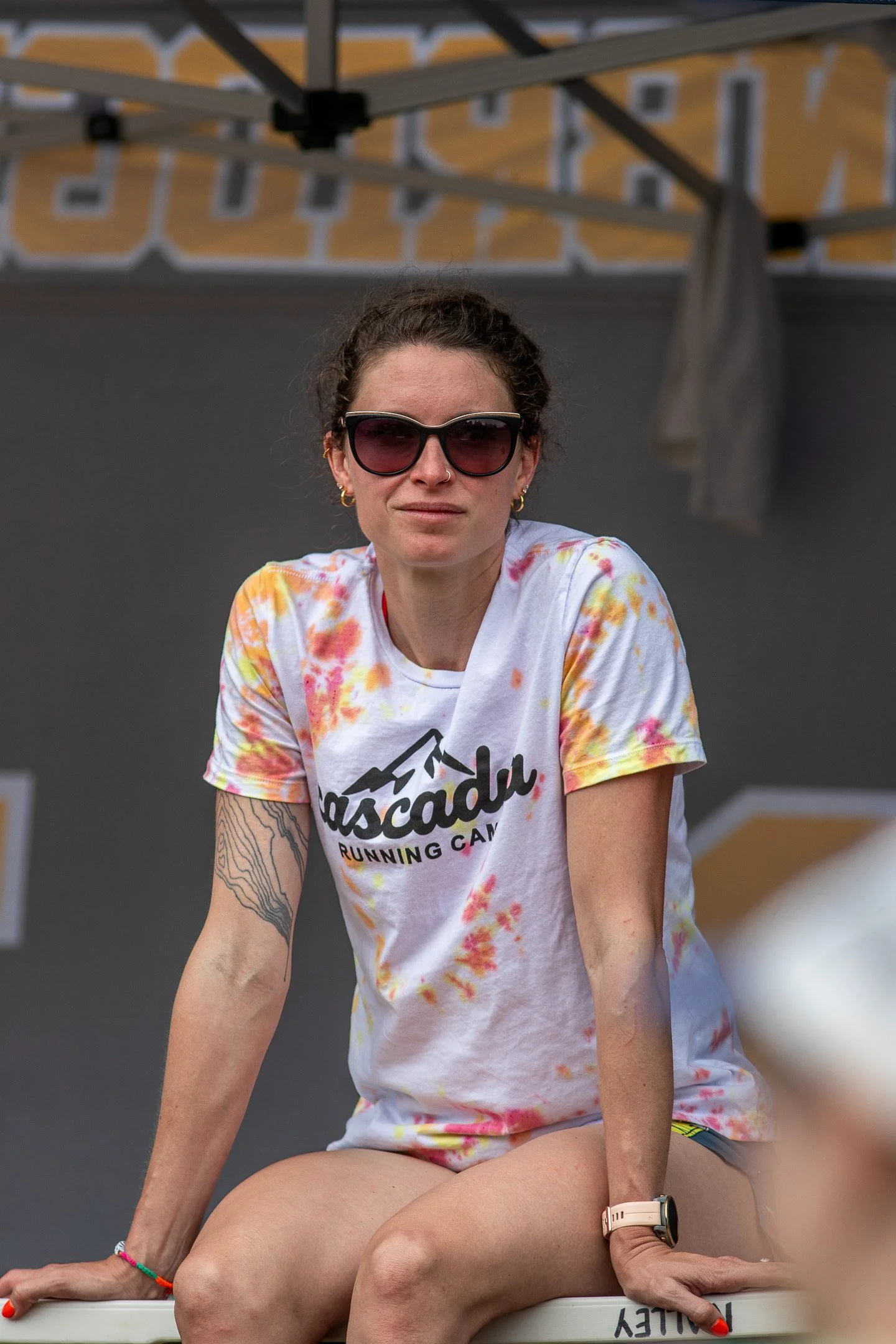 A woman with short brown hair wearing dark sunglasses, a white t-shirt with colorful tie-dye patterns, and various jewelry, sitting on a ledge outdoors.