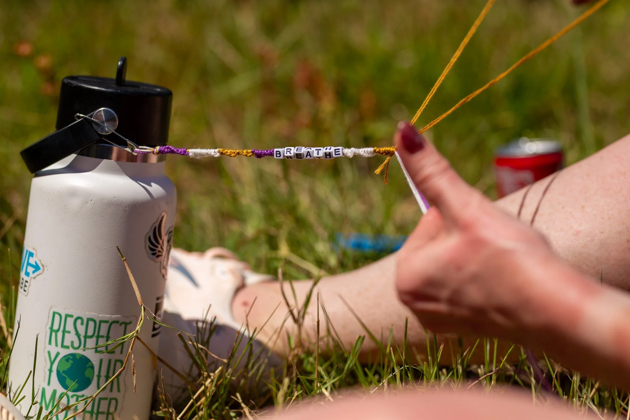 Close-up of a hand holding bead string with the words 'BREATHE' and 'RESPECT' on it, attached to a bottle, outdoors on grass.