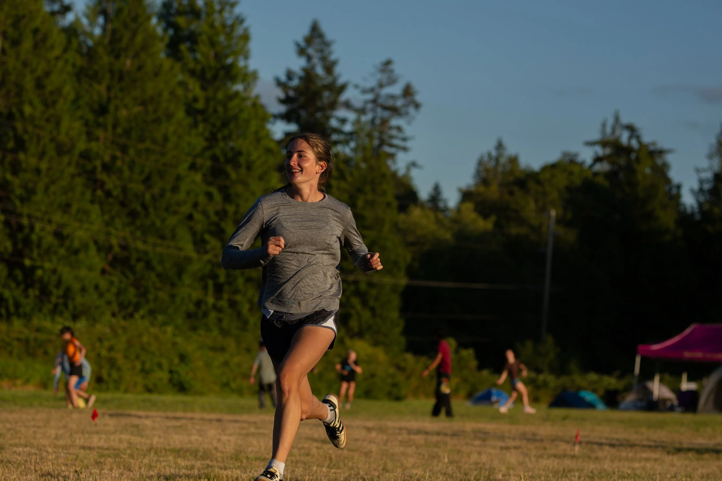 A woman running outdoors on a grassy field during sunset, with trees and other people in the background.