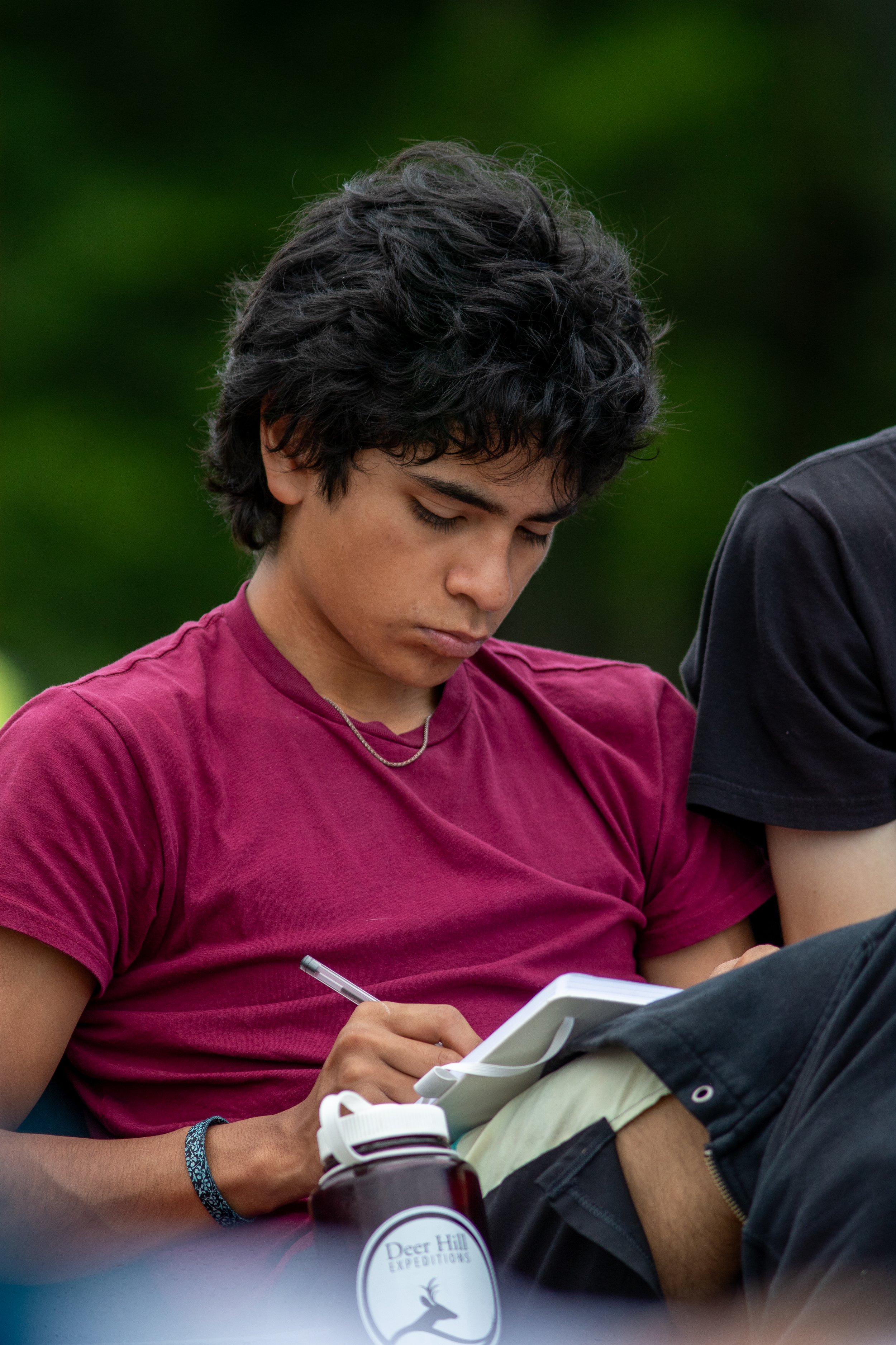 A young person with dark, curly hair wearing a maroon t-shirt, sitting outdoors with a notebook and pen, surrounded by greenery.