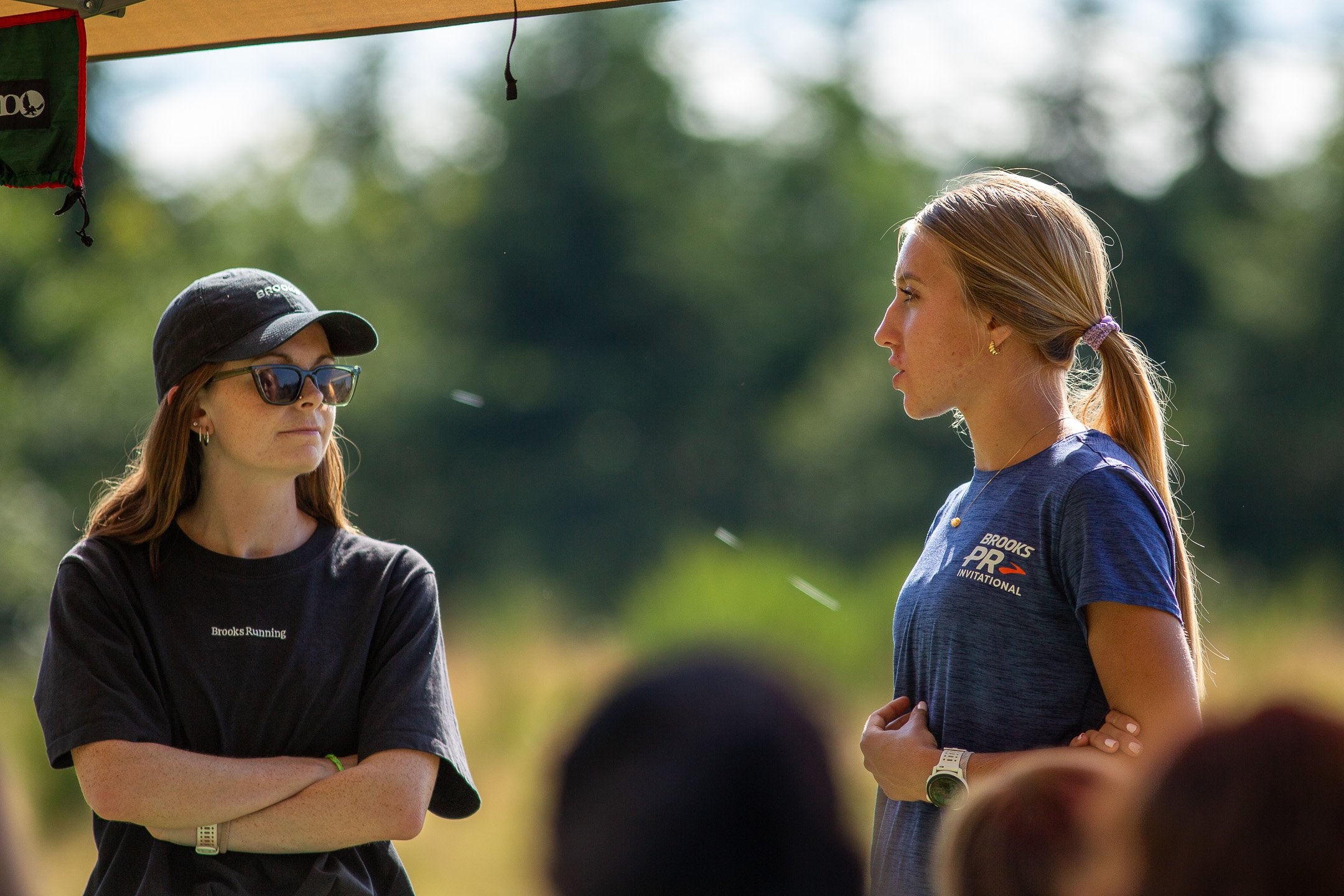 Two young women with long hair, one with glasses and a black cap, and the other with hair tied back, wearing a blue athletic shirt, standing outdoors under a canopy, engaged in conversation with a blurred background of trees.