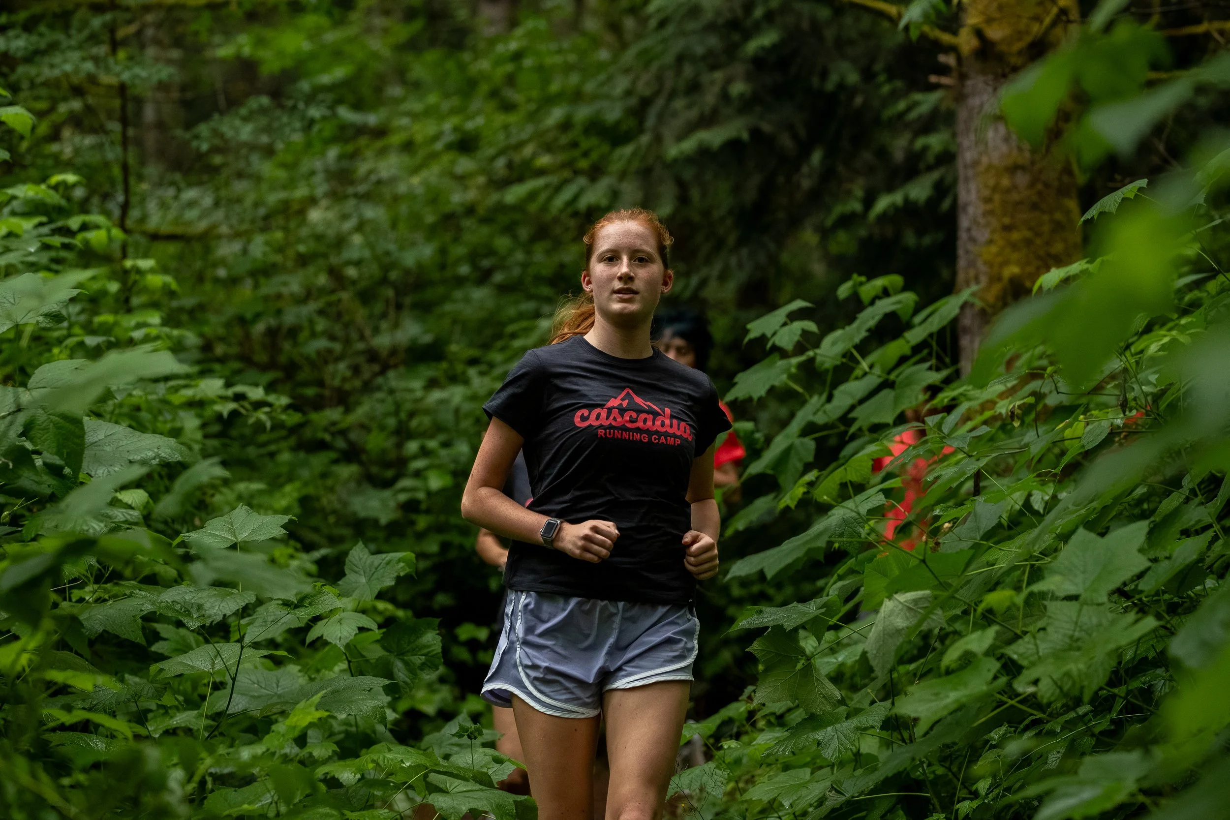 A young woman with red hair running through a lush green forest, wearing a black t-shirt with a red mountain logo and gray shorts.