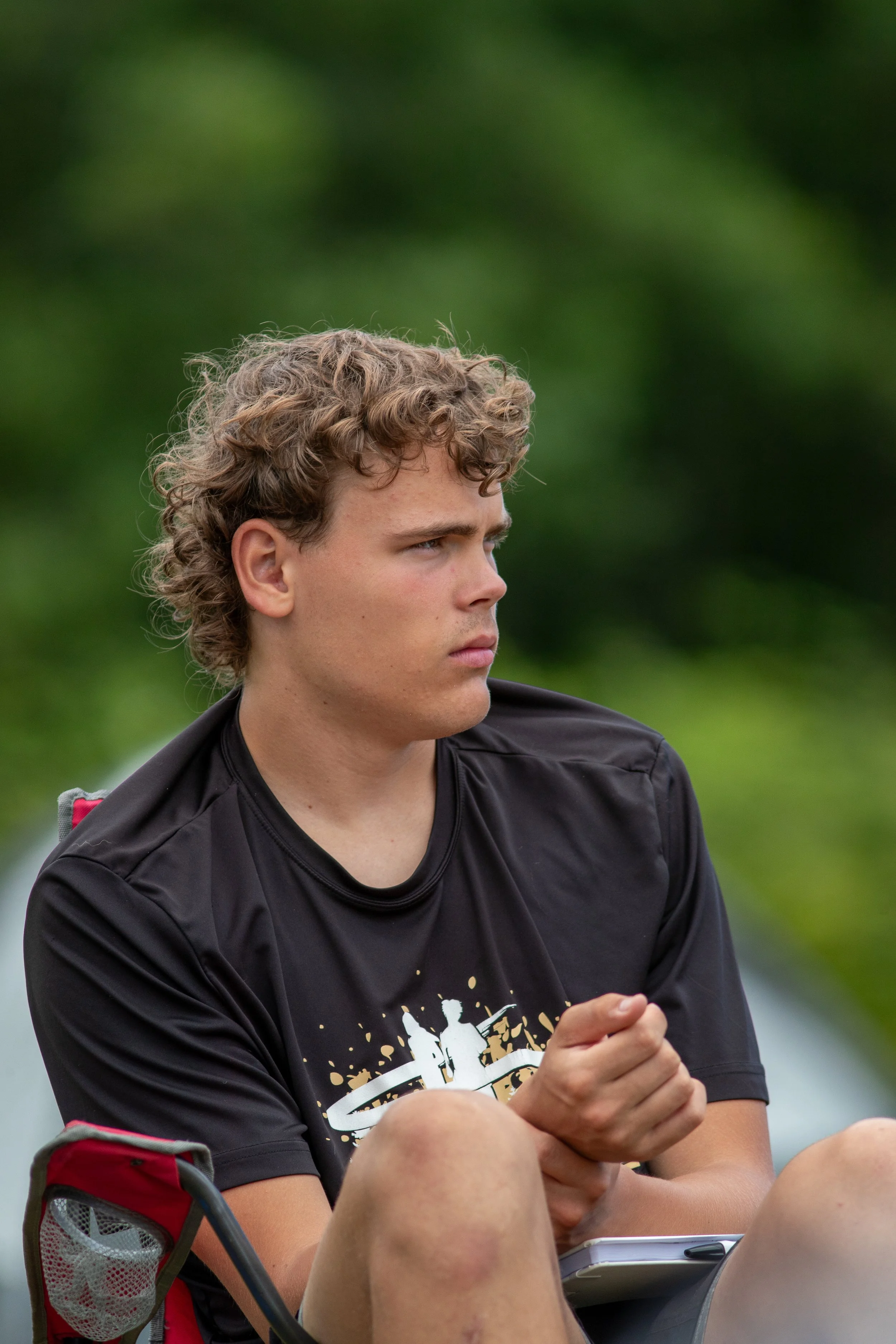 A young man with curly blond hair sitting outdoors, wearing a black graphic T-shirt, with a serious expression, and a green background.