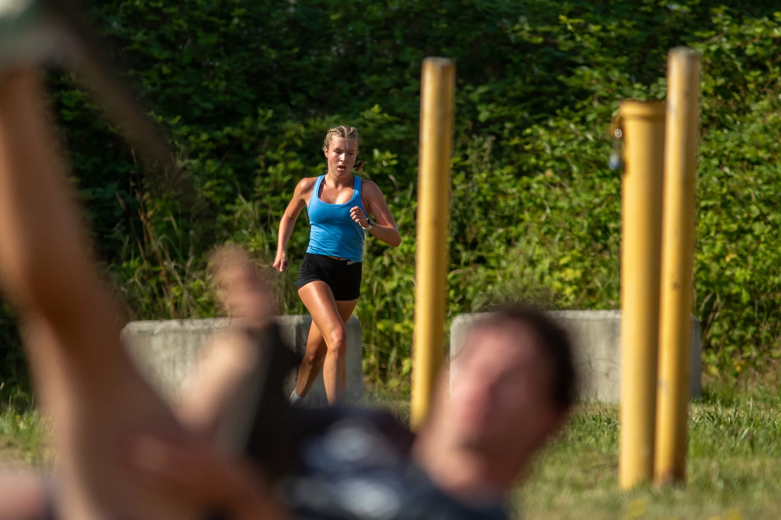A young woman running outdoors next to yellow poles and greenery, wearing a blue tank top and black shorts.