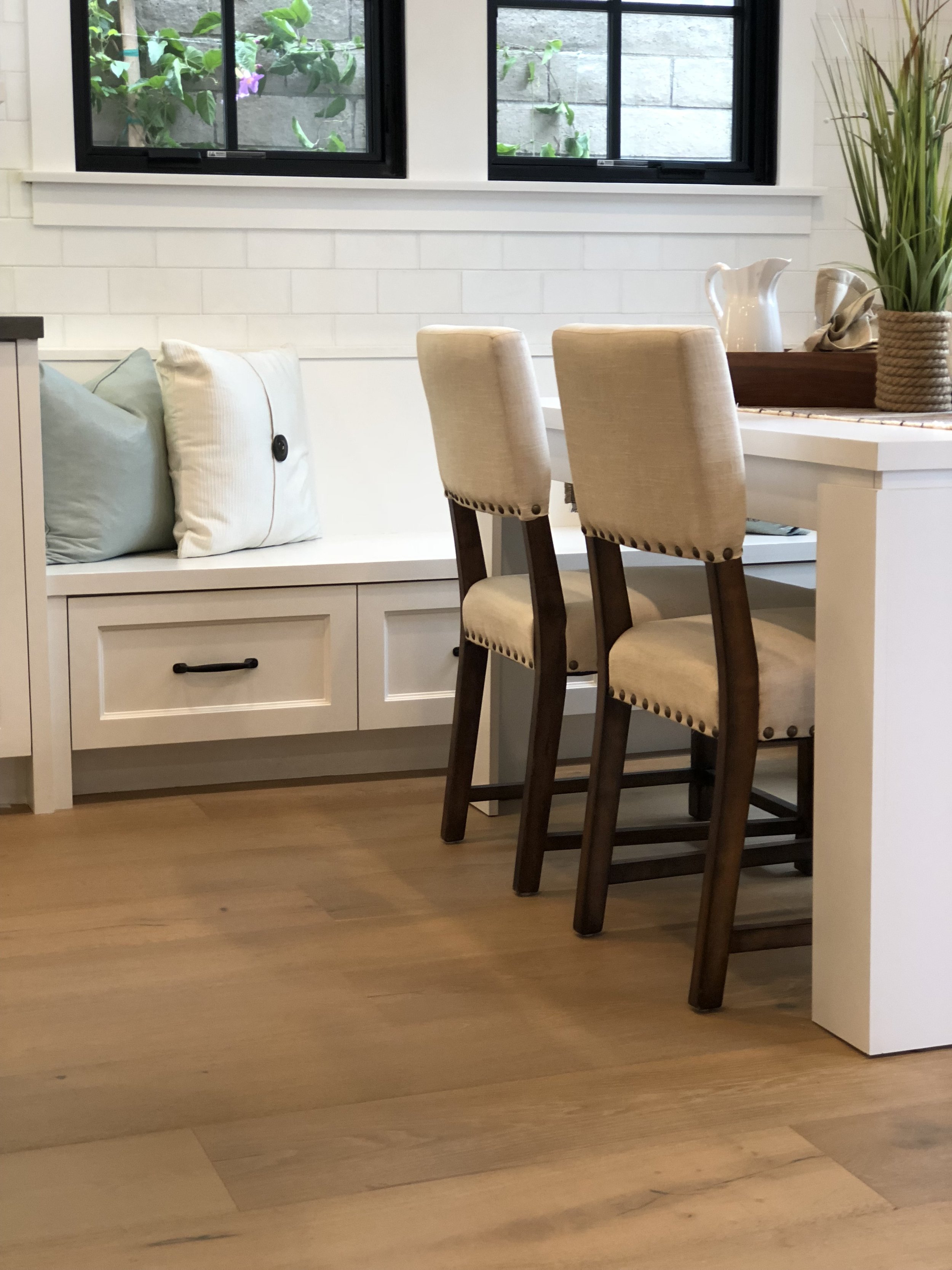 A cozy kitchen nook with a white built-in bench under two black framed windows, decorated with pillows. Two beige upholstered chairs with wooden frames are placed at a white dining table, which has a tray with a large plant, pitchers, and a napkin on