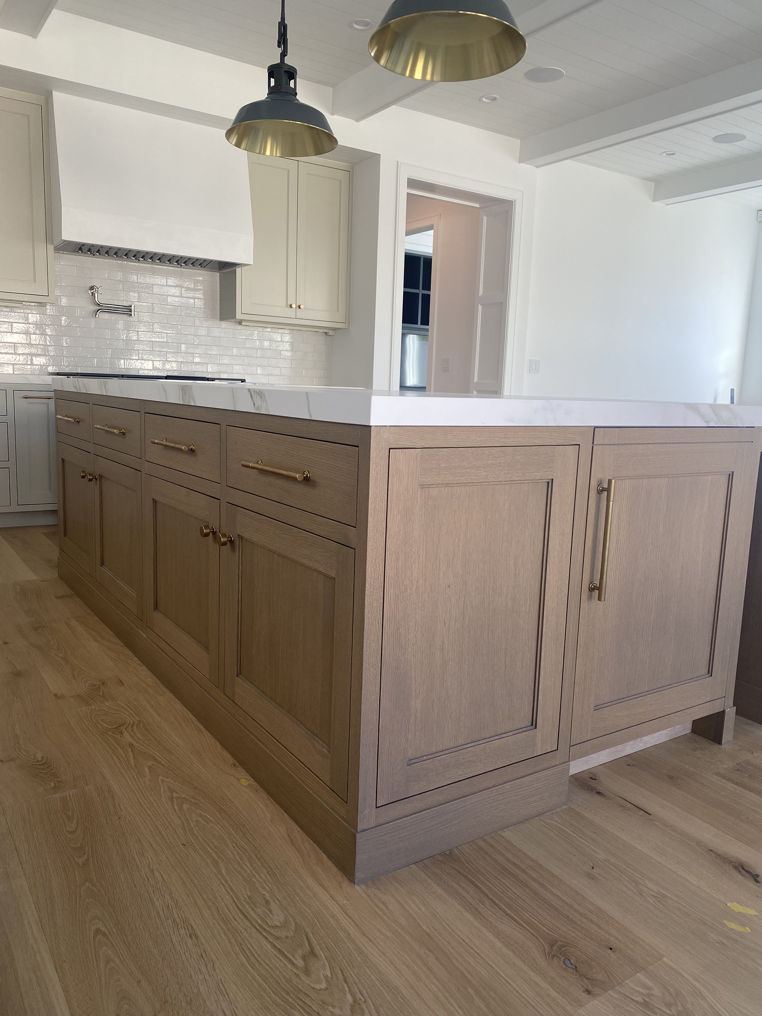 Wooden kitchen island with brass handles, white marble countertop, light-colored flooring, and modern pendant lights.