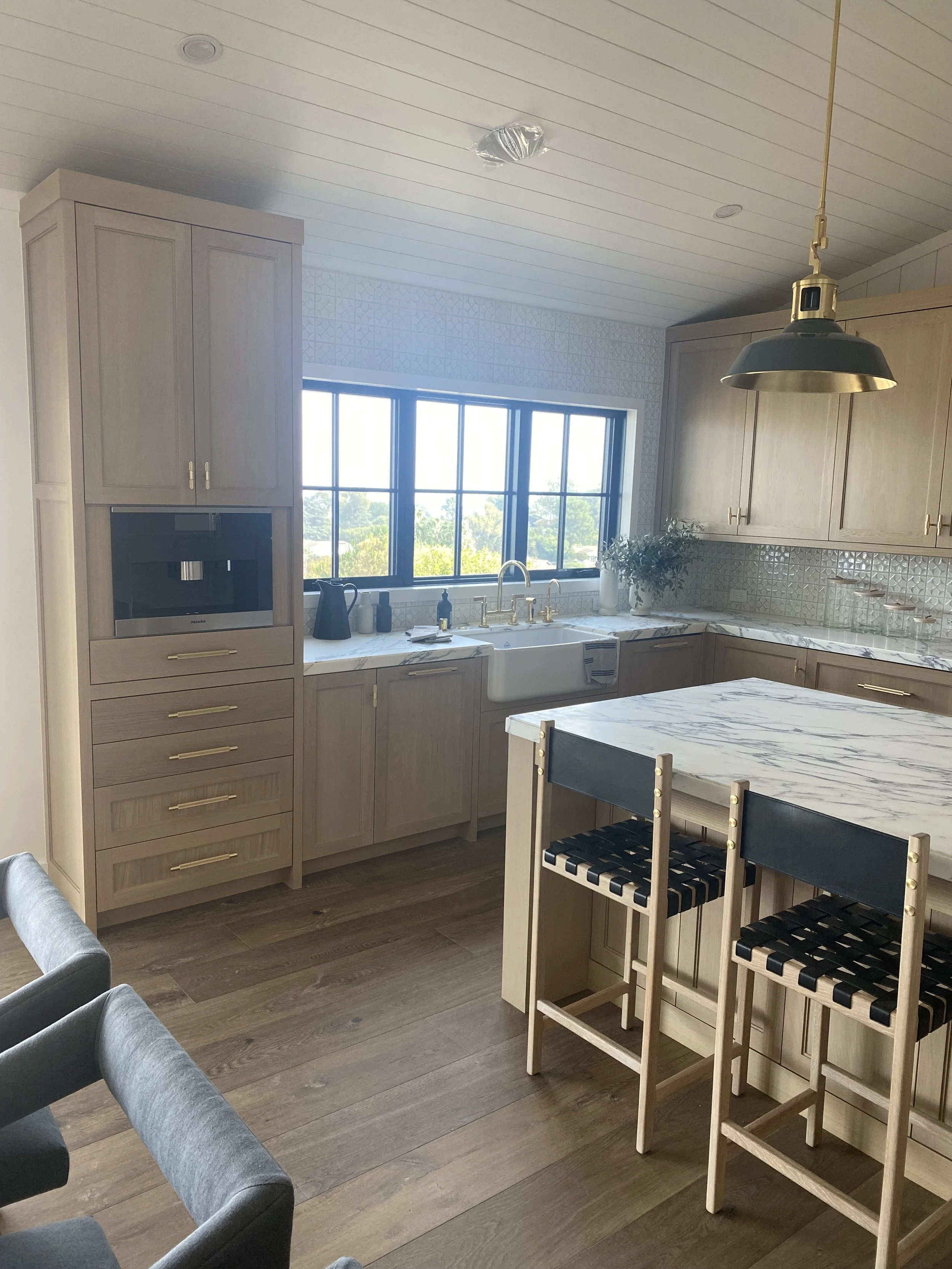 A bright, modern kitchen with light wood cabinets, a large window over the sink, marble countertops, and two black-and-wood bar stools at a marble-topped island.