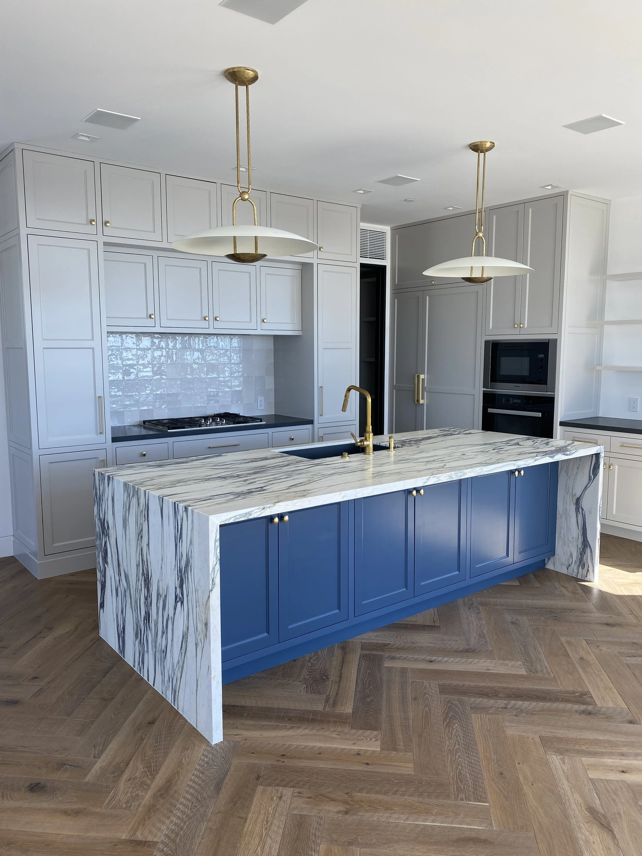 Modern kitchen with white cabinetry, blue island with marble top, gold hardware, and gold fixtures, including a faucet and pendant lights.