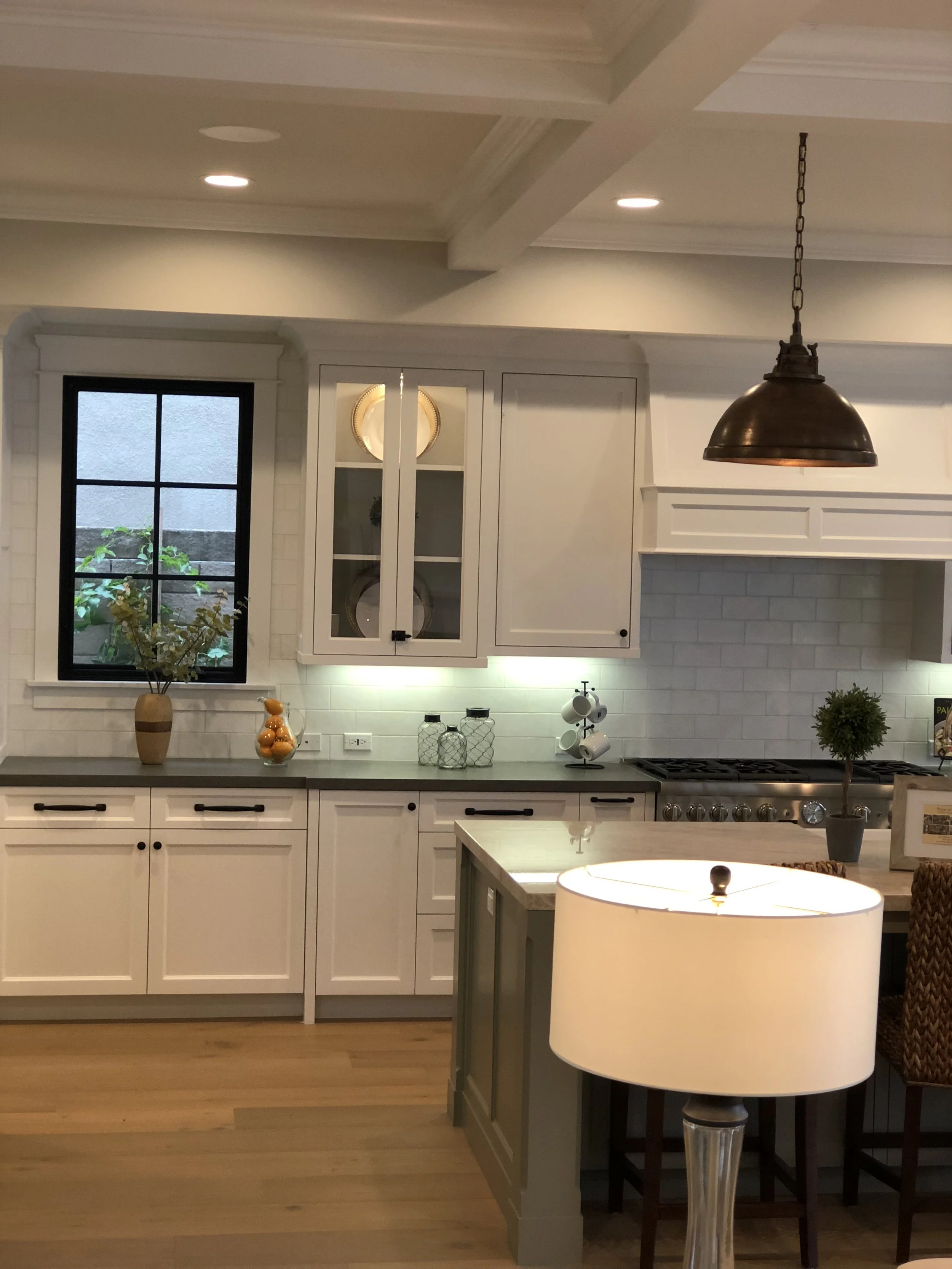 A modern kitchen with white cabinets, a gray countertop, and a window with a view of greenery. Decor includes a vase with flowers, a bowl of oranges, glass jars, a small potted topiary on the island, and a hanging copper pendant light. There's also a