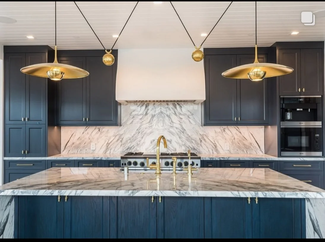 Modern kitchen with navy blue cabinets, marble countertops, and gold fixtures, including pendant lights and faucet.