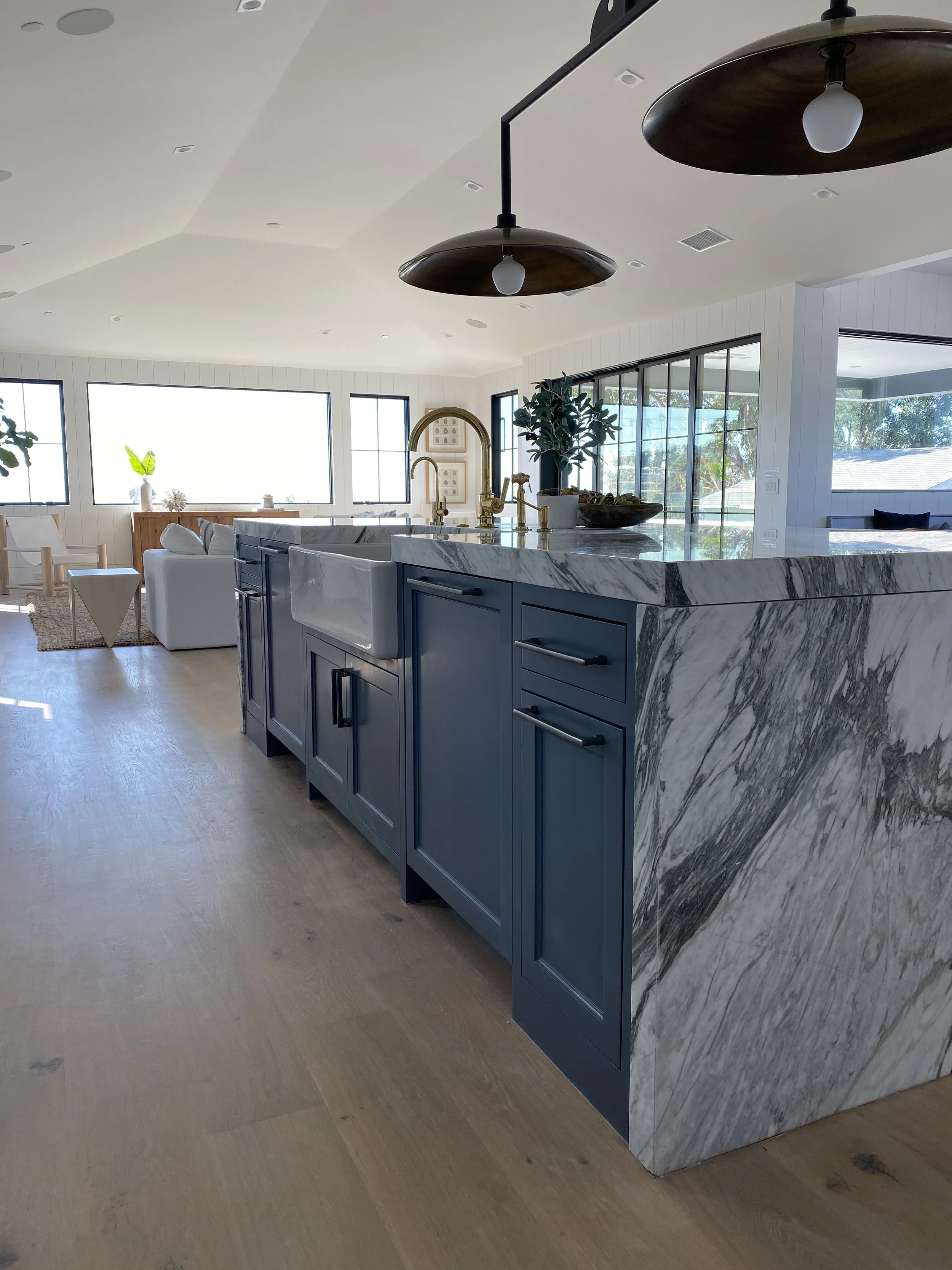 Modern open-concept kitchen with a blue island, marble countertops, gold fixtures, and large windows providing natural light.