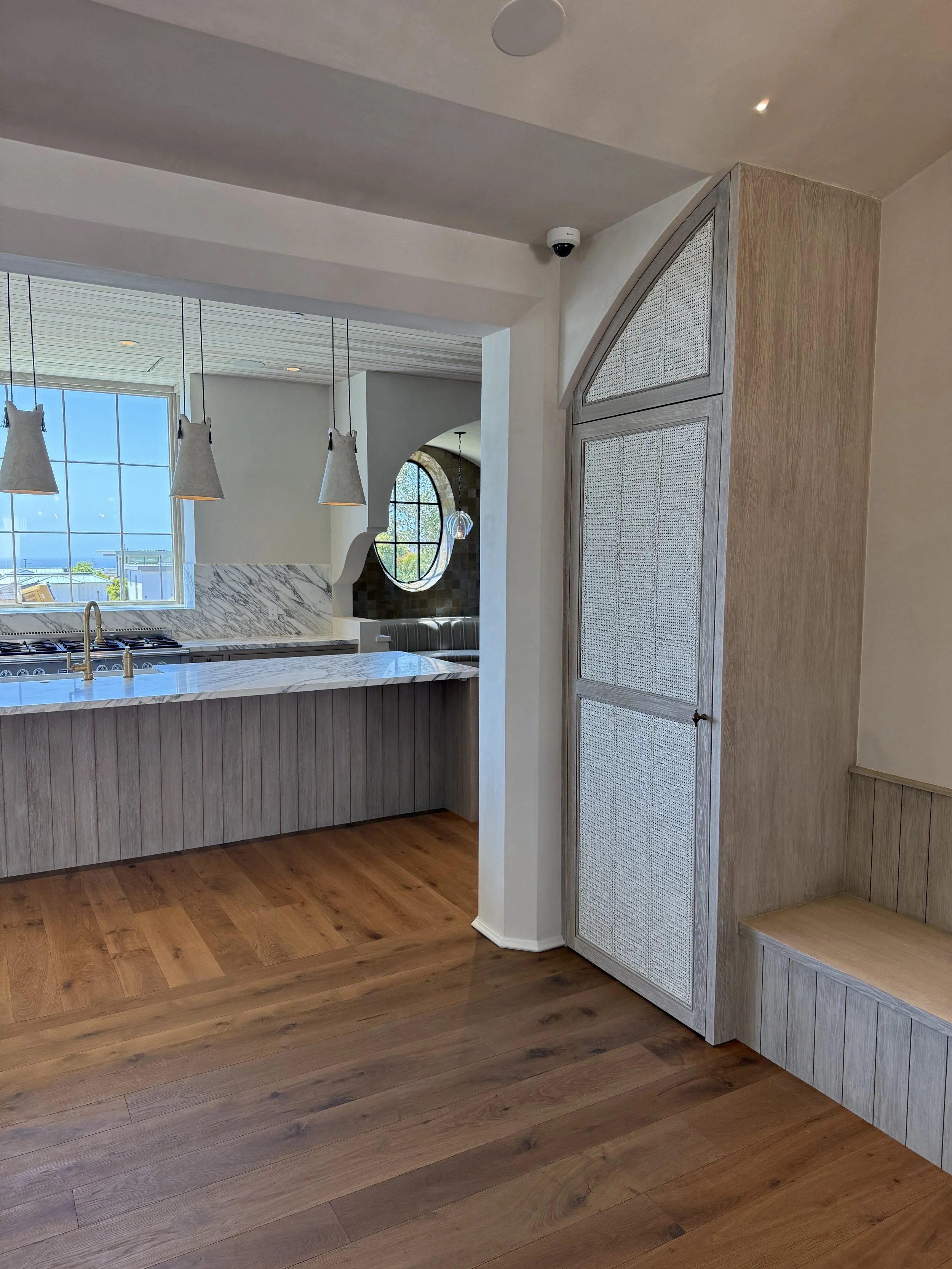 Interior view of a modern kitchen with hanging pendant lights, marble countertop, large window, and wooden floors.