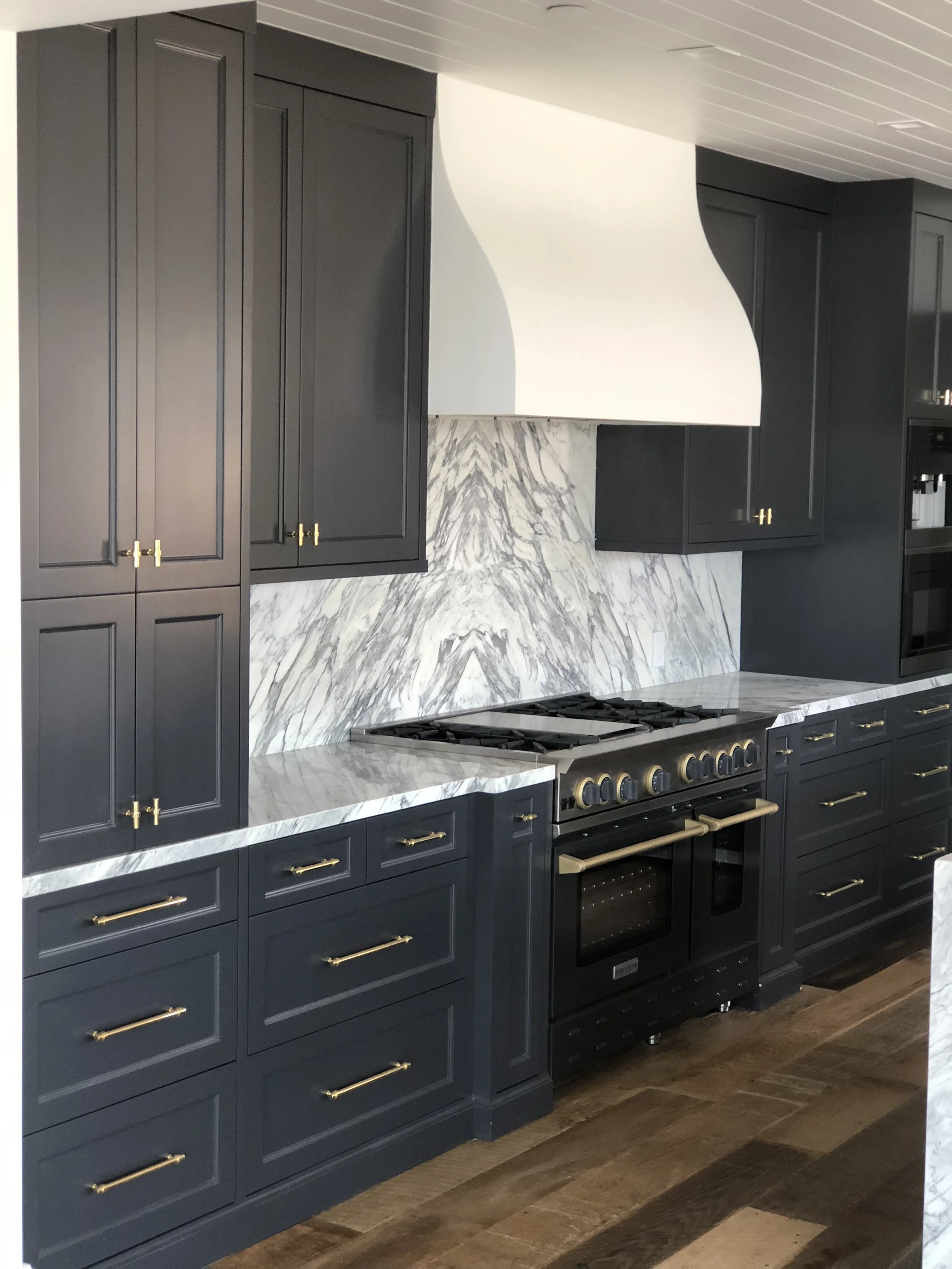 Kitchen featuring navy blue cabinets with gold handles, marble countertops, and a marble backsplash with gray veining, a white range hood, and a black stove.
