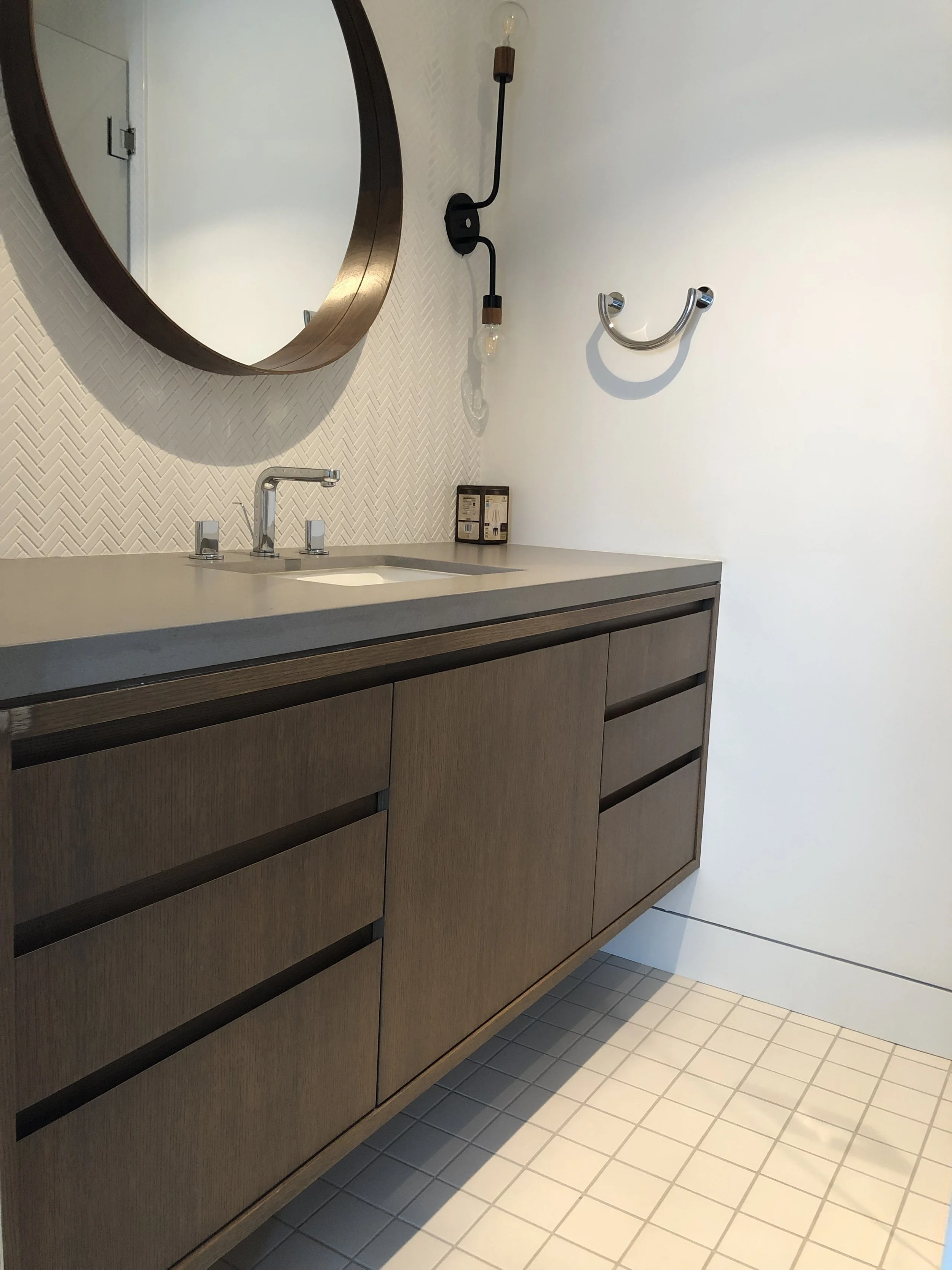Modern bathroom vanity with a round mirror, faucet, and a small shelf. Wall-mounted light fixture and towel hook. Beige tiled floor and textured white wall.