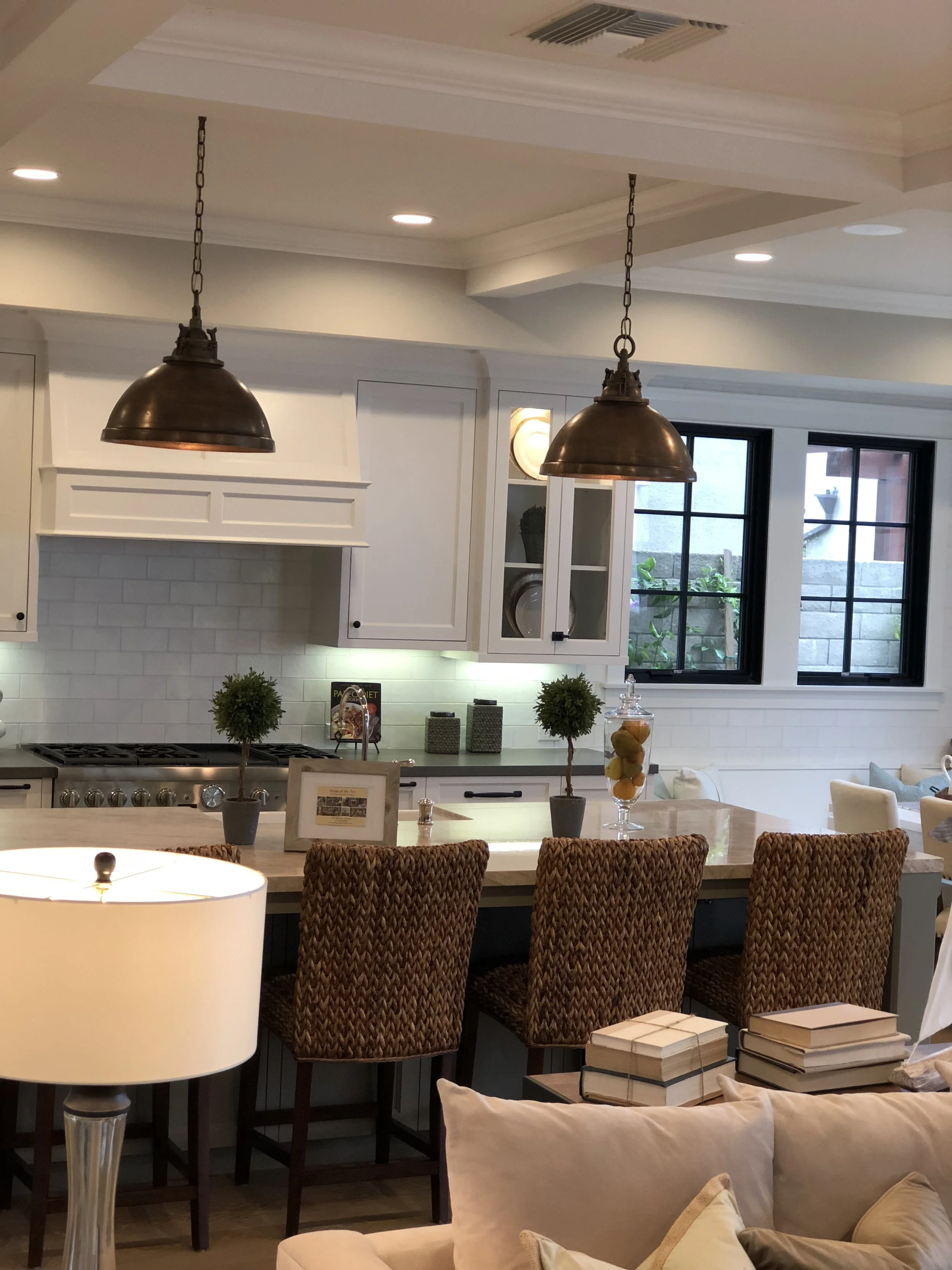 Modern kitchen with white cabinets, black-framed windows, pendant lights, and a marble island with woven barstools. Decor includes small trees, framed photo, books, and a glass jar with yellow lemons.