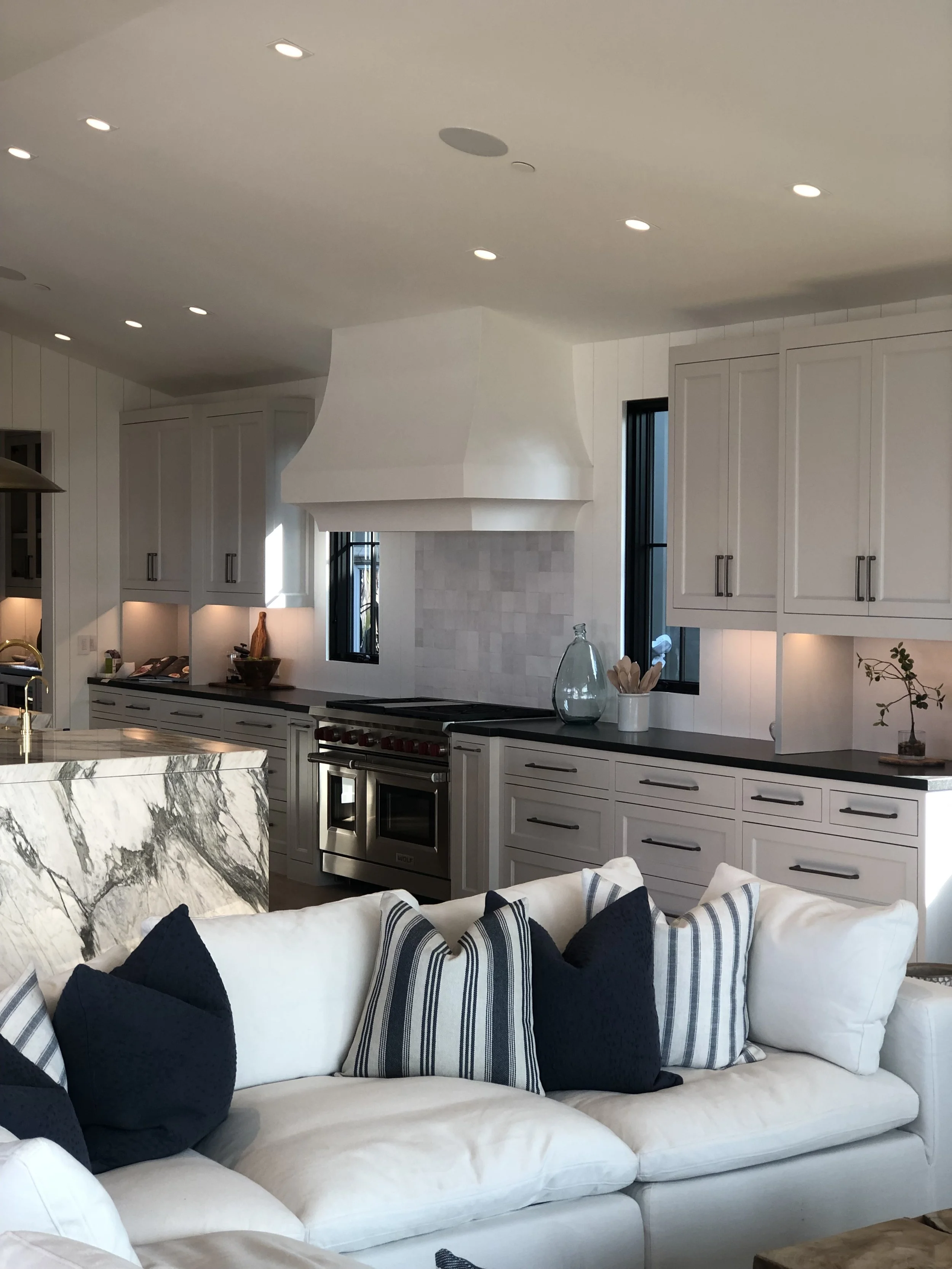 Modern kitchen with white cabinets, a marble island, a range with a hood, and a white sofa with striped and dark blue pillows in the foreground.