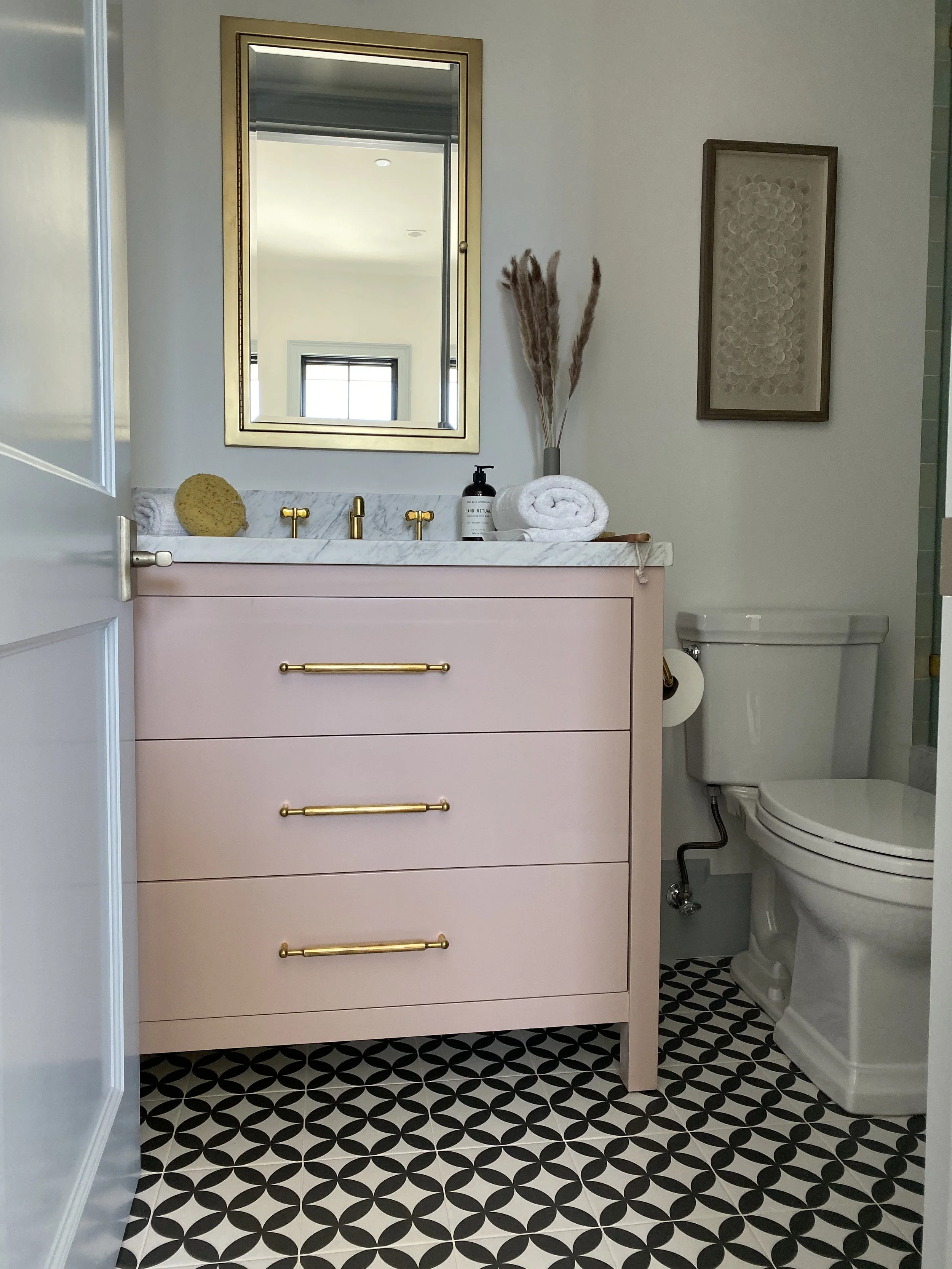 A modern bathroom featuring a pink vanity with gold handles, a white marble countertop, a large mirror with a gold frame, and decorative items such as a soap sponge, a soap dispenser, rolled towel, and a vase with dried pampas grass. To the right, th