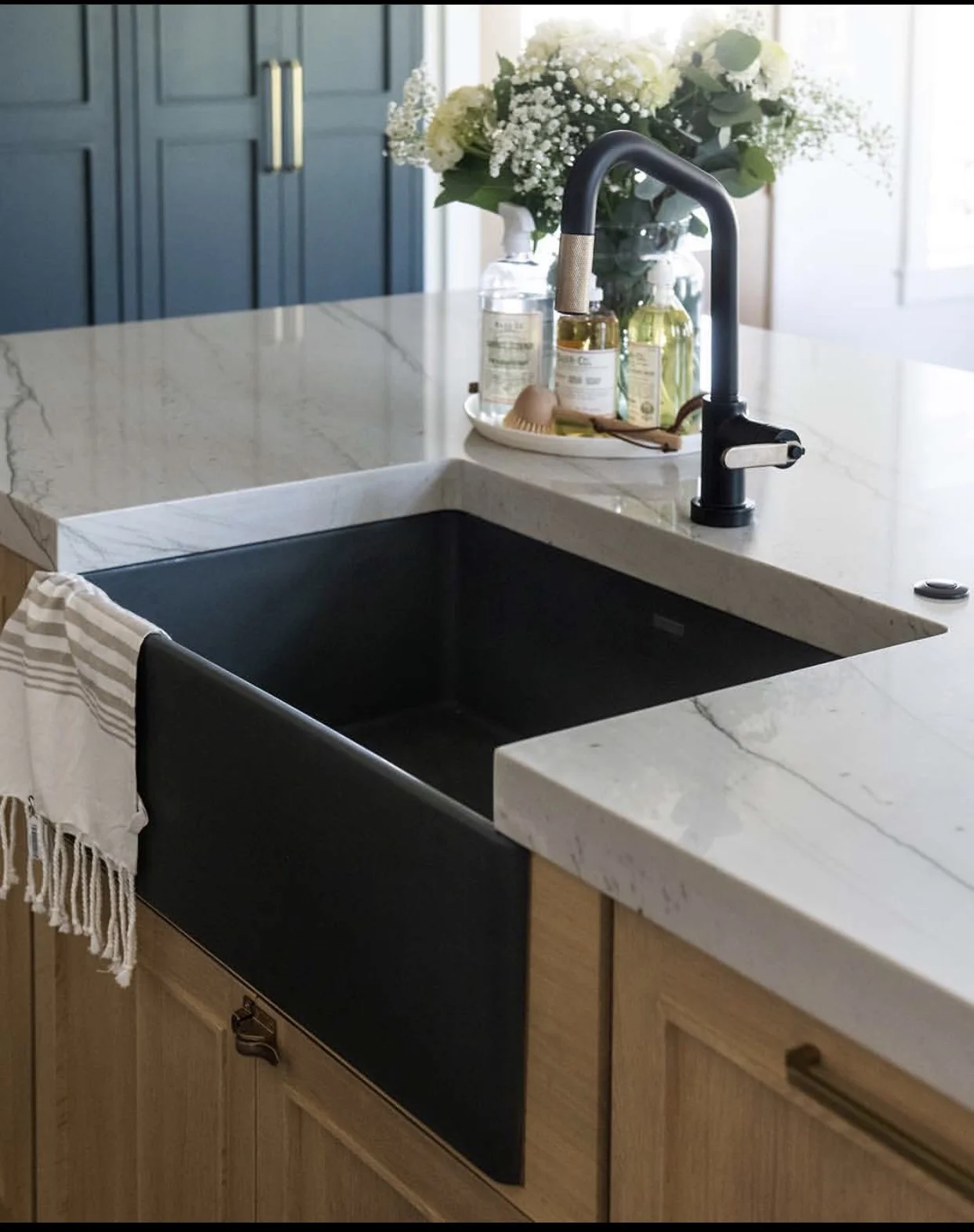 A kitchen countertop with a black sink, a black faucet, a white marble surface, a drying towel, and a bouquet of white flowers in the background.
