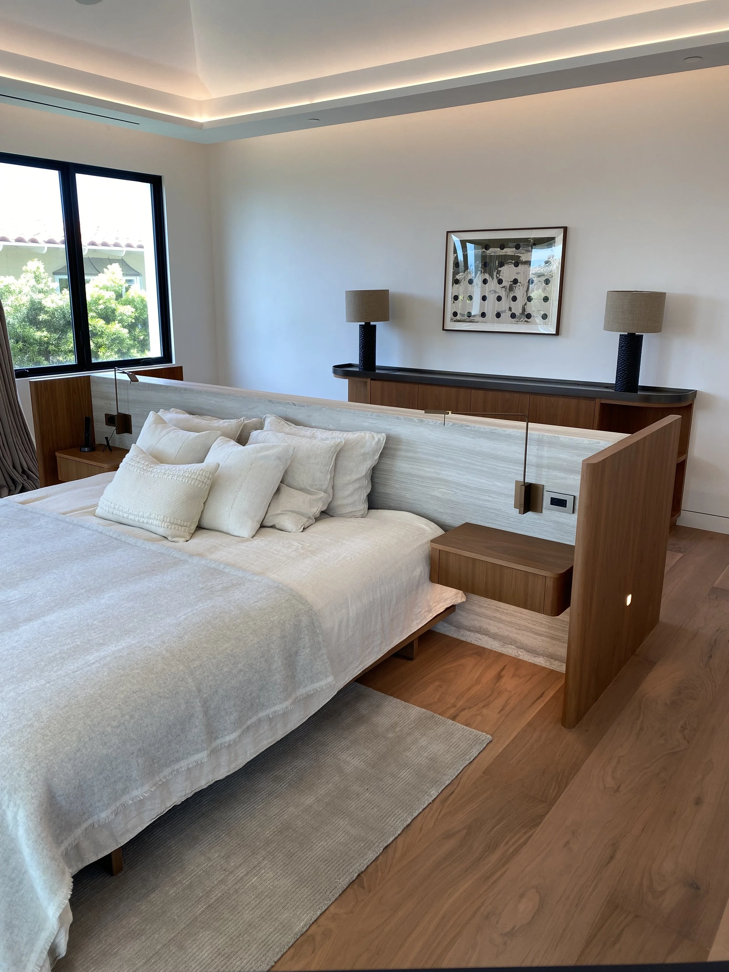 Modern bedroom with a wooden bed frame, white bedding, multiple pillows, wooden side tables, black table lamps with beige shades, a wooden dresser with a framed abstract art piece, a large window with curtains, and a wooden floor.