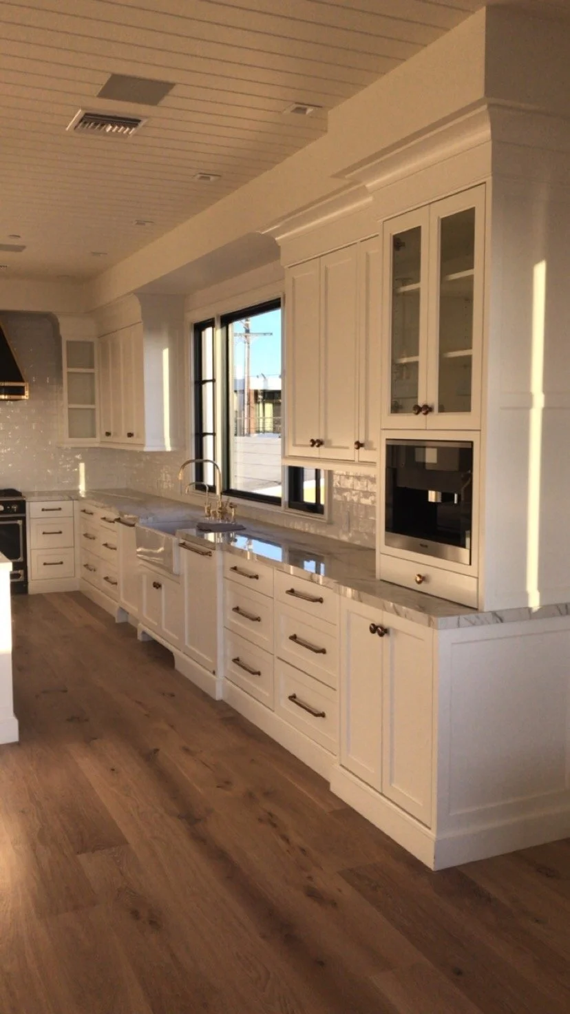 Modern kitchen with white cabinets, marble countertops, and hardwood flooring, sunlight shining through a large window.