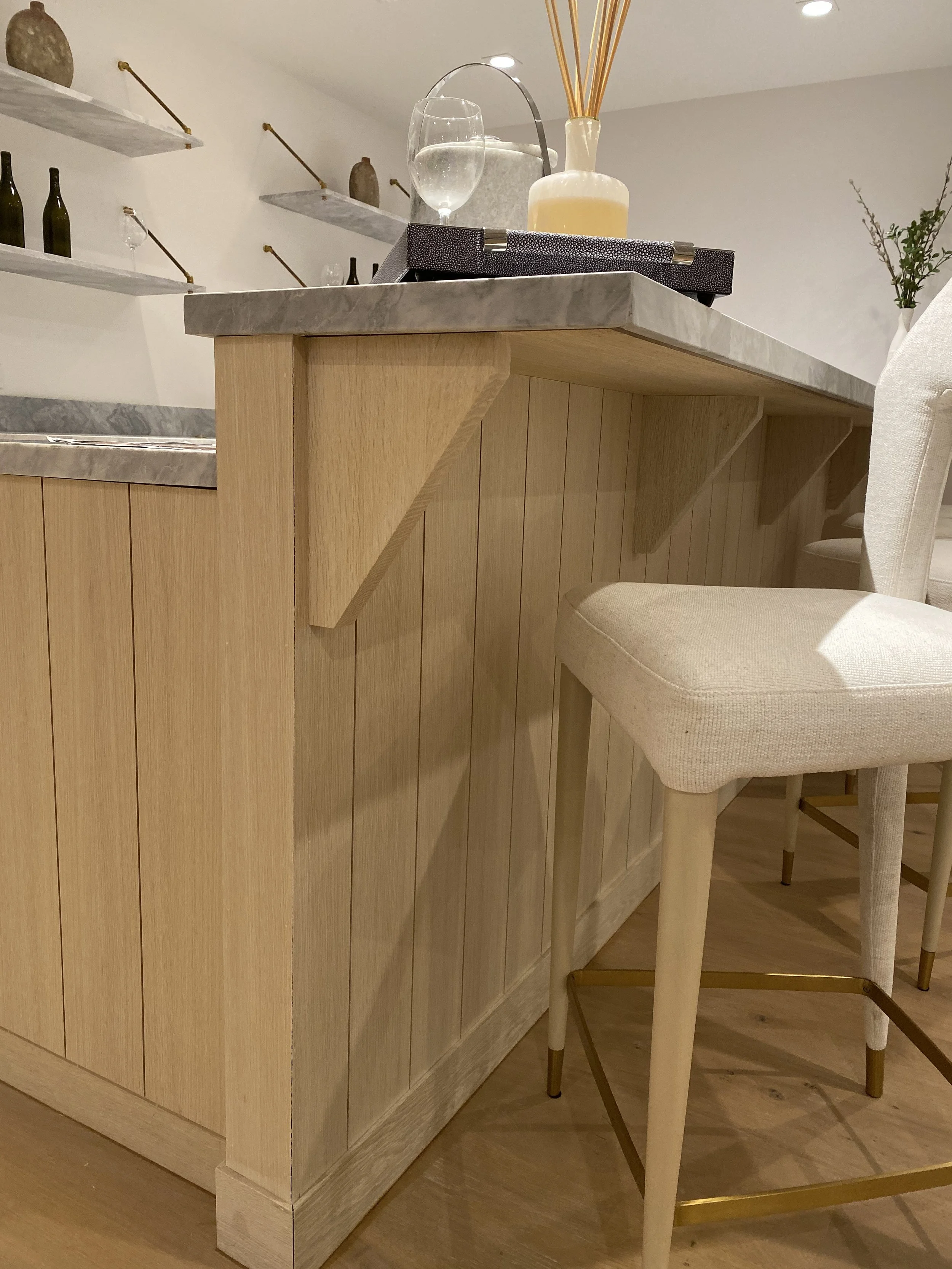 Close-up of a kitchen island with a marble countertop, a beige bar stool with gold accents, and decorative items on shelves in the background.