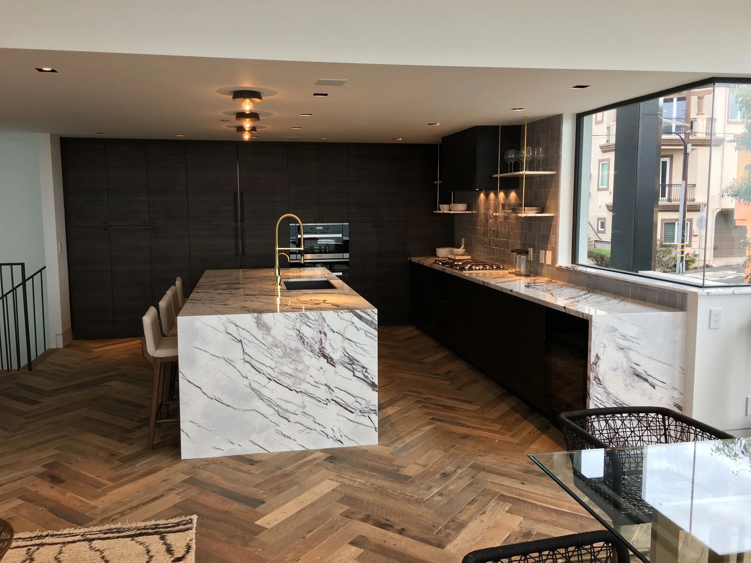 Modern kitchen with dark wood cabinets, marble island, gold faucet, and large window with city view.