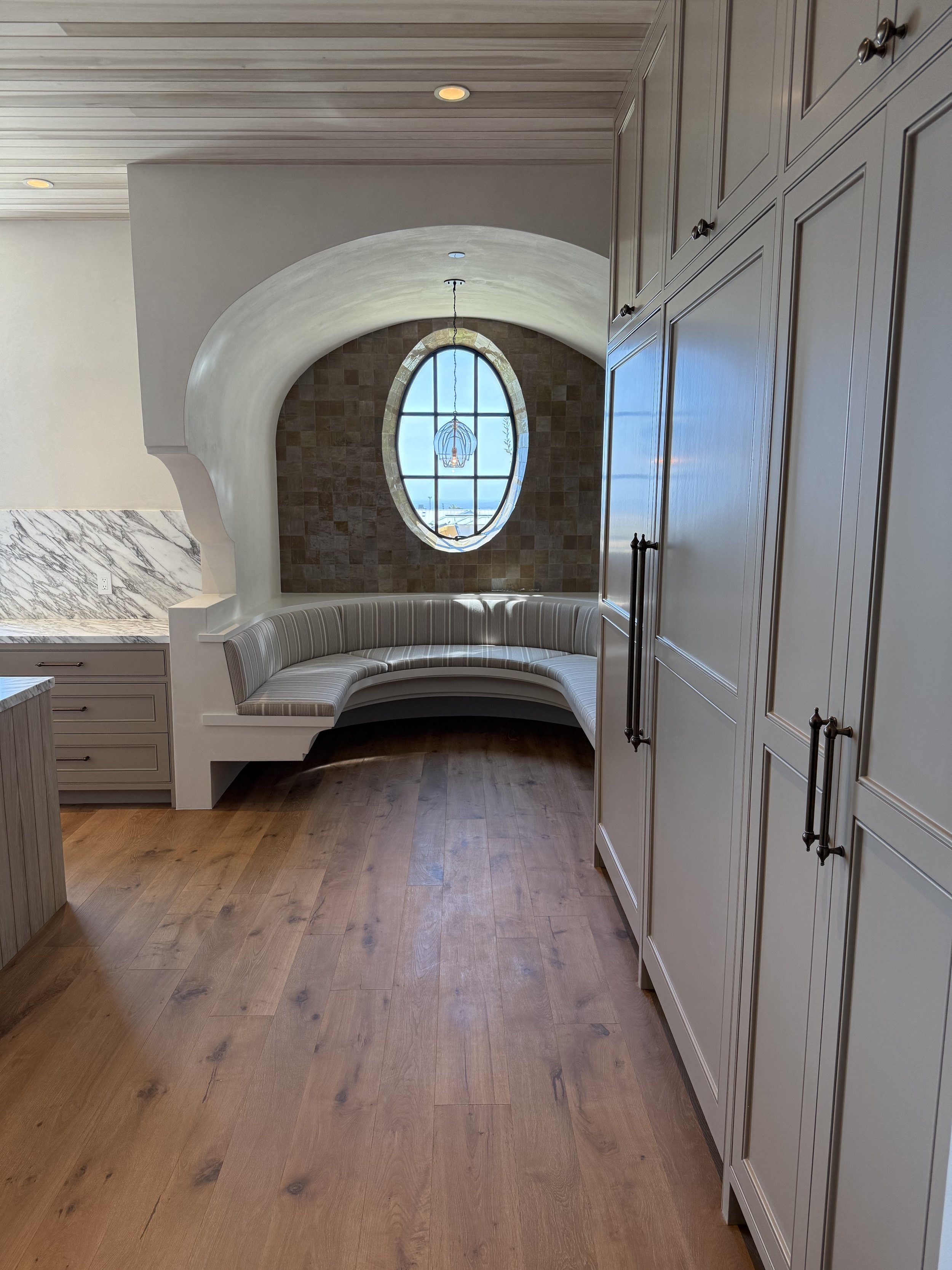 Kitchen nook with a curved built-in bench under an oval window with a view of the ocean, surrounded by tiled wall, with light wood flooring and cabinetry.