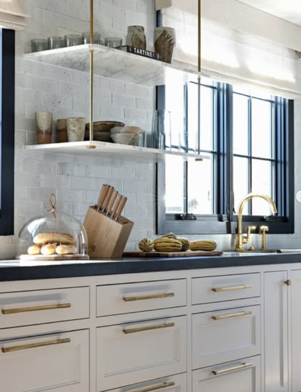 Modern kitchen with white cabinets, gold hardware, black countertop, open shelves holding dishes and glasses, a window with dark frames, and a gold faucet.