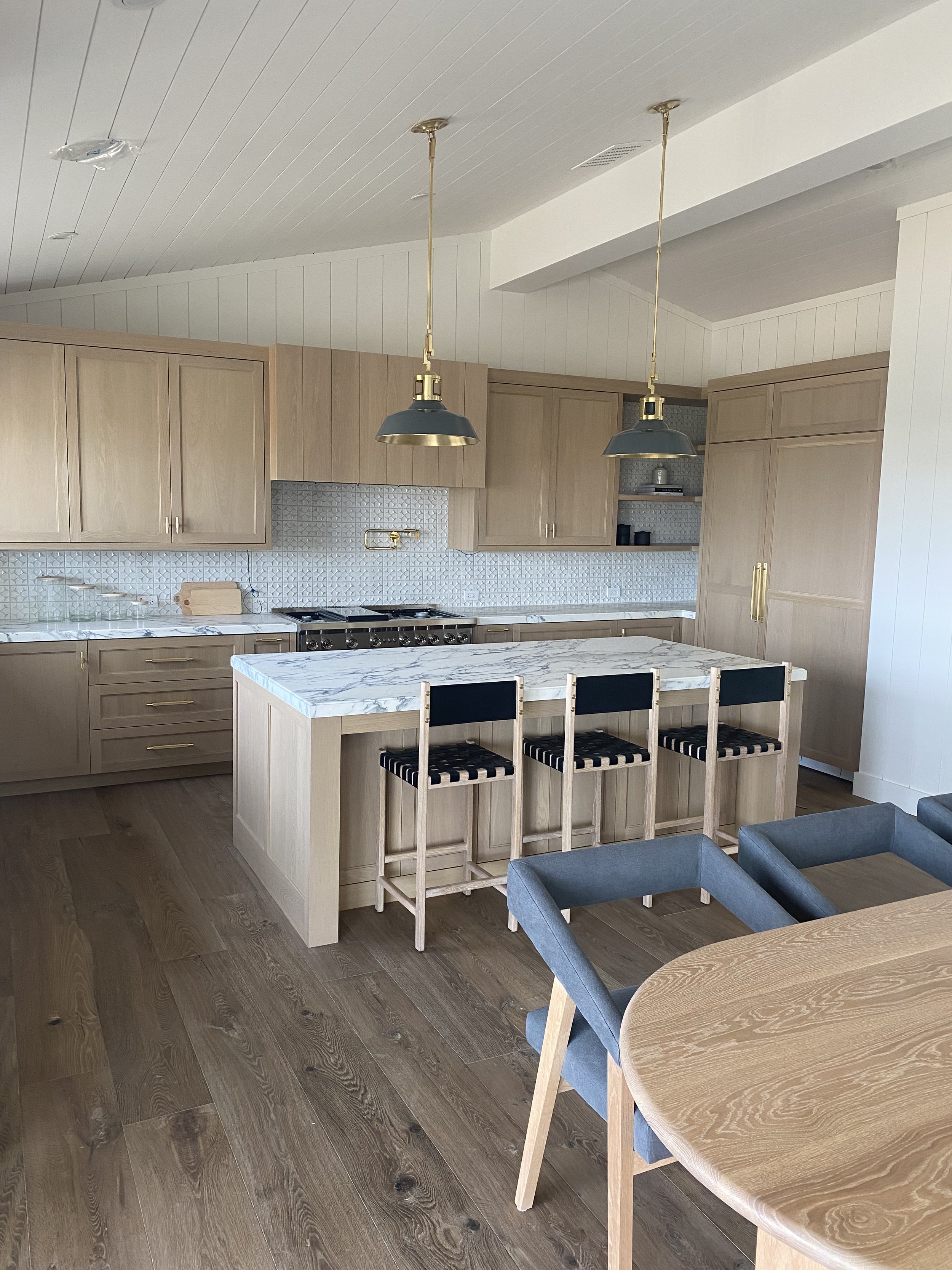 Modern kitchen with light wood cabinets, marble island, blue pendant lights, patterned backsplash, and wooden chairs, with part of dining table visible.