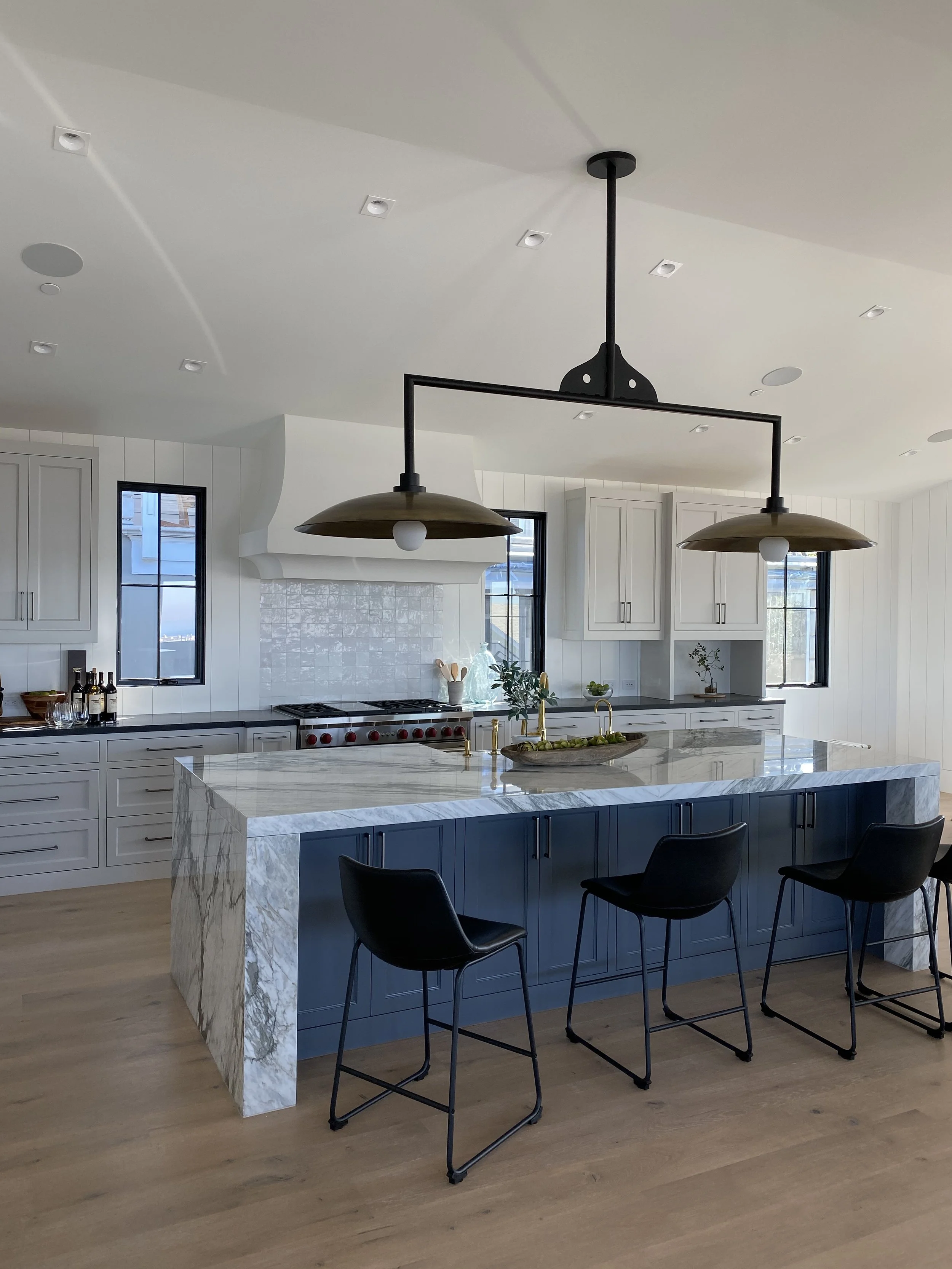 Modern kitchen with white cabinets, a marble island with blue base, black bar stools, black-framed windows, and a dual pendant light fixture.