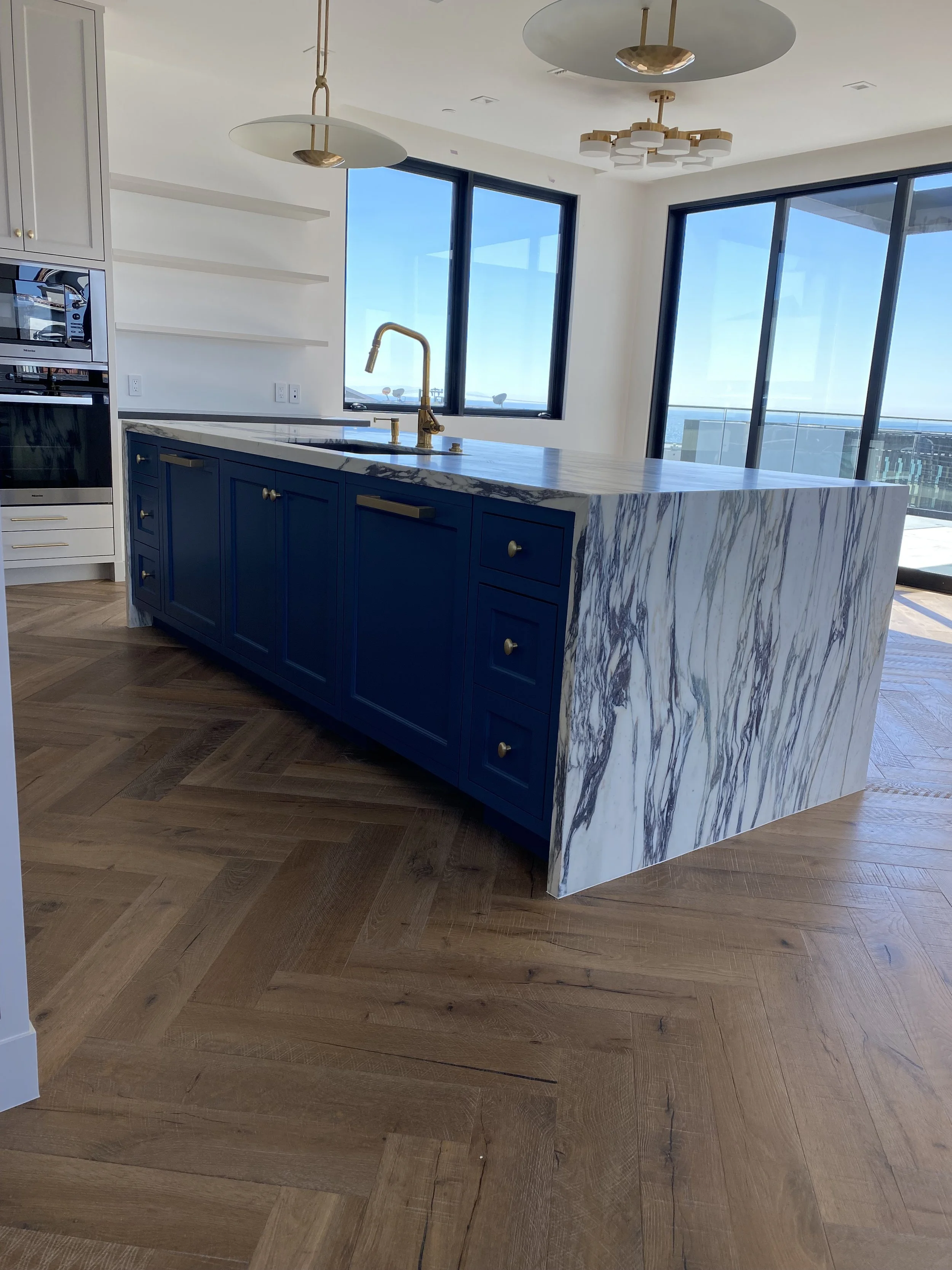 Modern kitchen island with blue cabinets and a marble countertop, gold fixtures, and large windows with a city view.