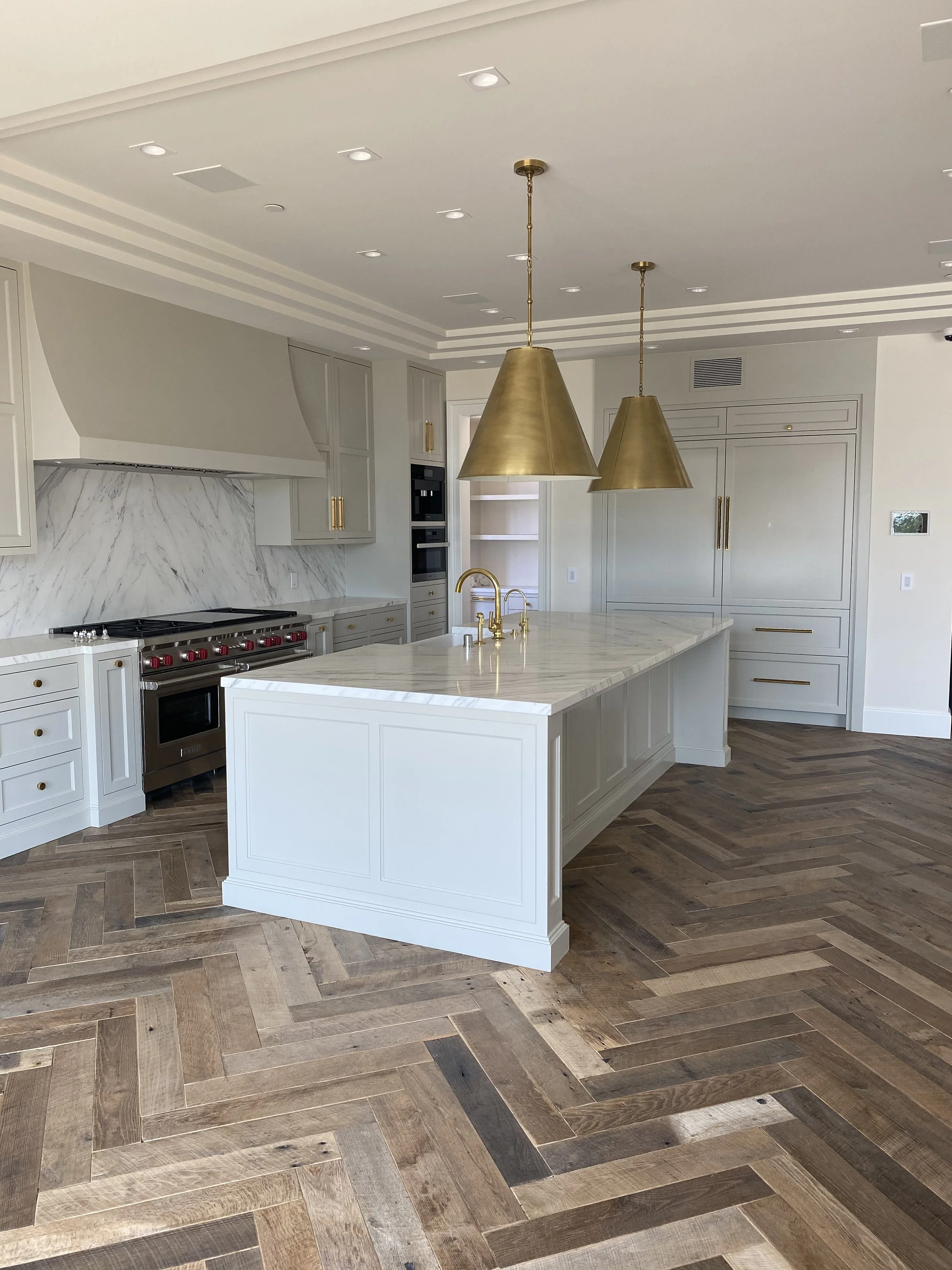 Modern kitchen with white cabinets, marble backsplash, large island with marble countertop, gold fixtures, and two large gold pendant lights, hardwood herringbone floor.