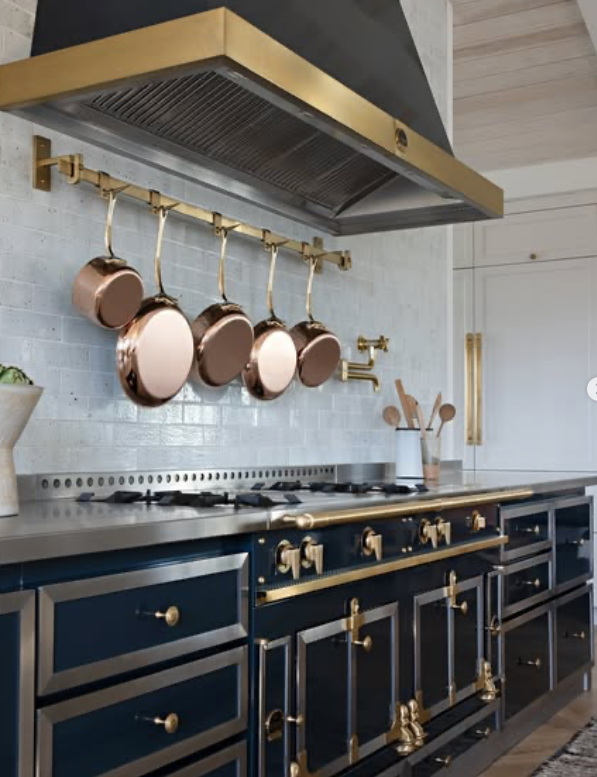 A modern kitchen with navy blue cabinets, gold hardware, a silver countertop, a white brick backsplash, and hanging copper pots over a stove with a gold range hood.