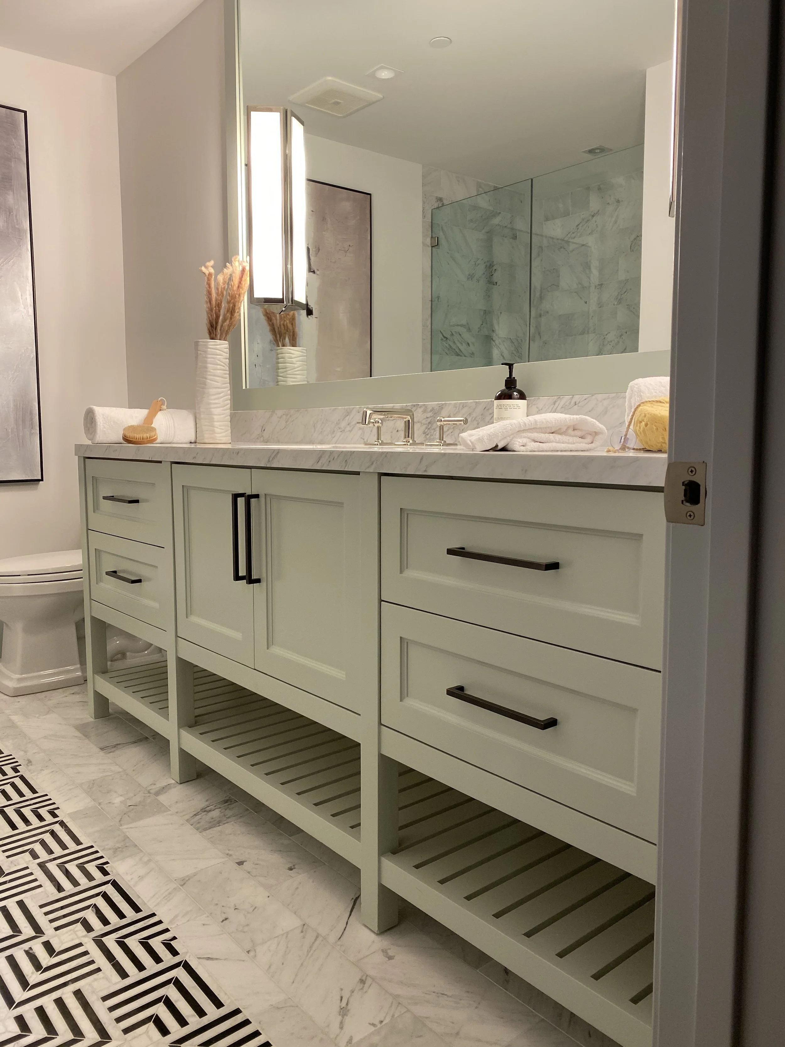 A bathroom vanity with a marble countertop, towels, a black soap dispenser, and decorative vases with dried pampas grass. A large mirror is above the vanity, and there is a walk-in shower with marble walls in the background.