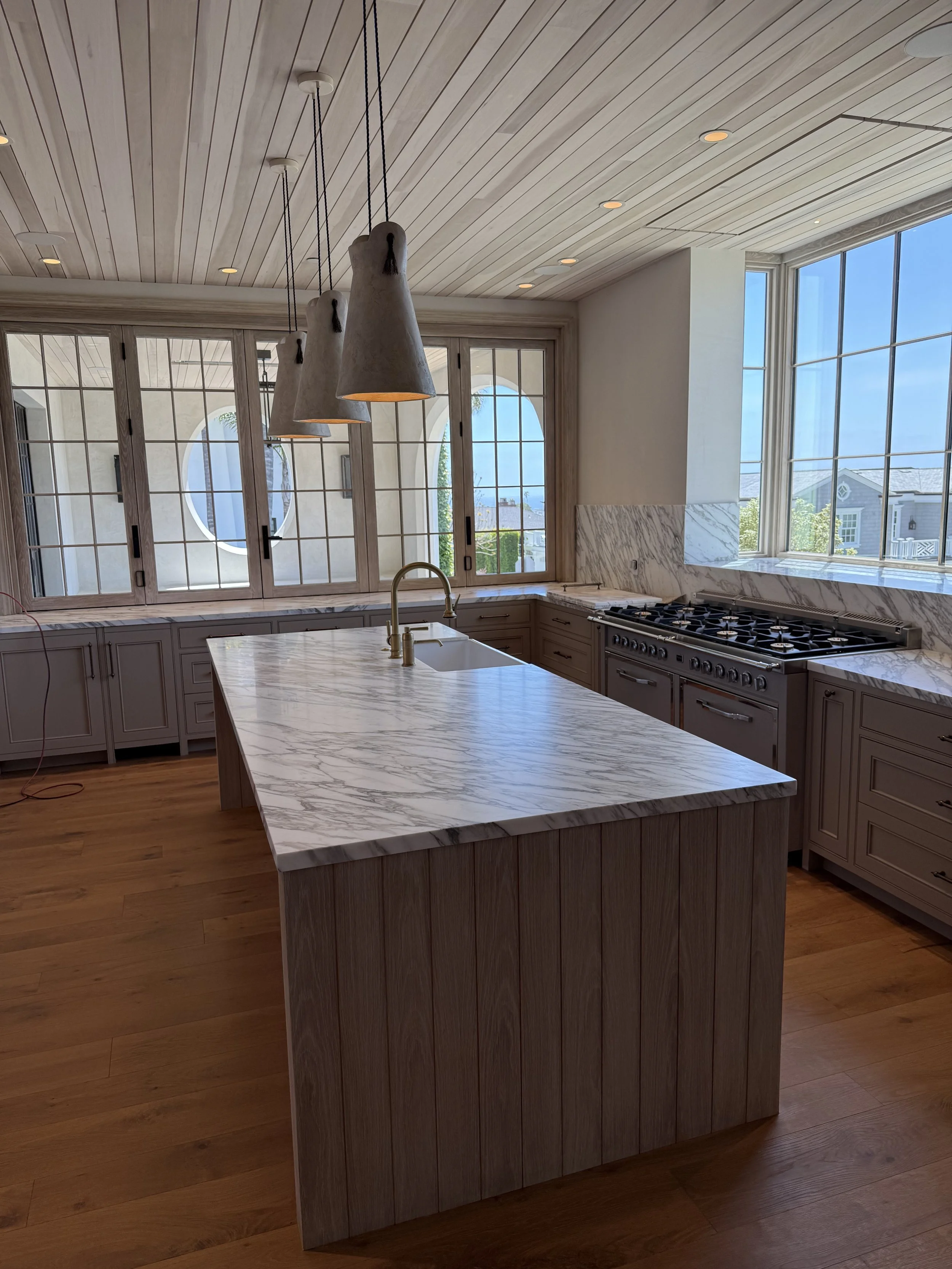 Modern kitchen with white marble island, large windows, wood flooring, pendant lights, and cabinetry.