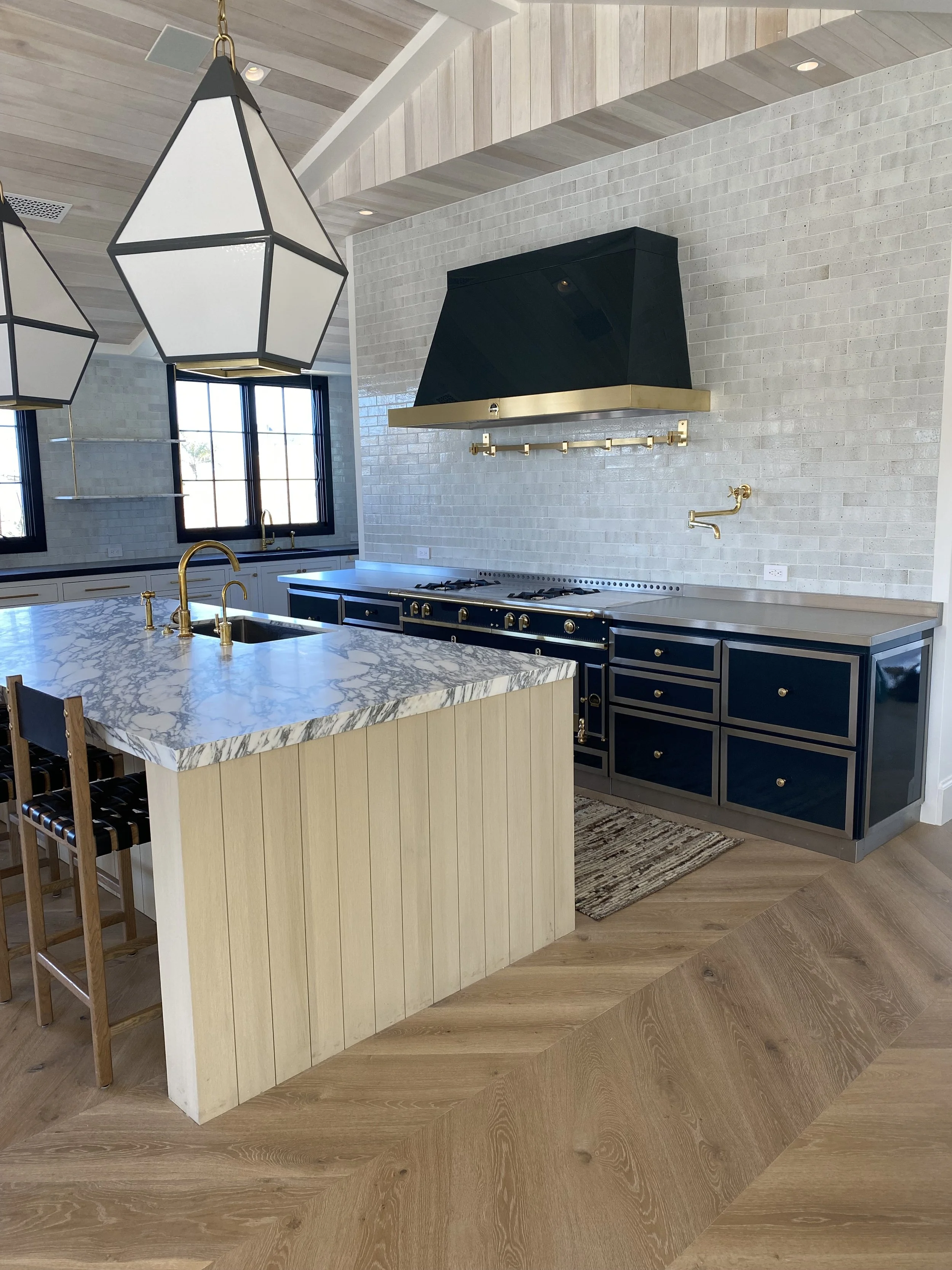Modern kitchen with black and gold accents, a marble island with a gold faucet, black cabinets with gold hardware, a black range hood with gold trim, white brick backsplash, pendant lights, and wooden flooring.