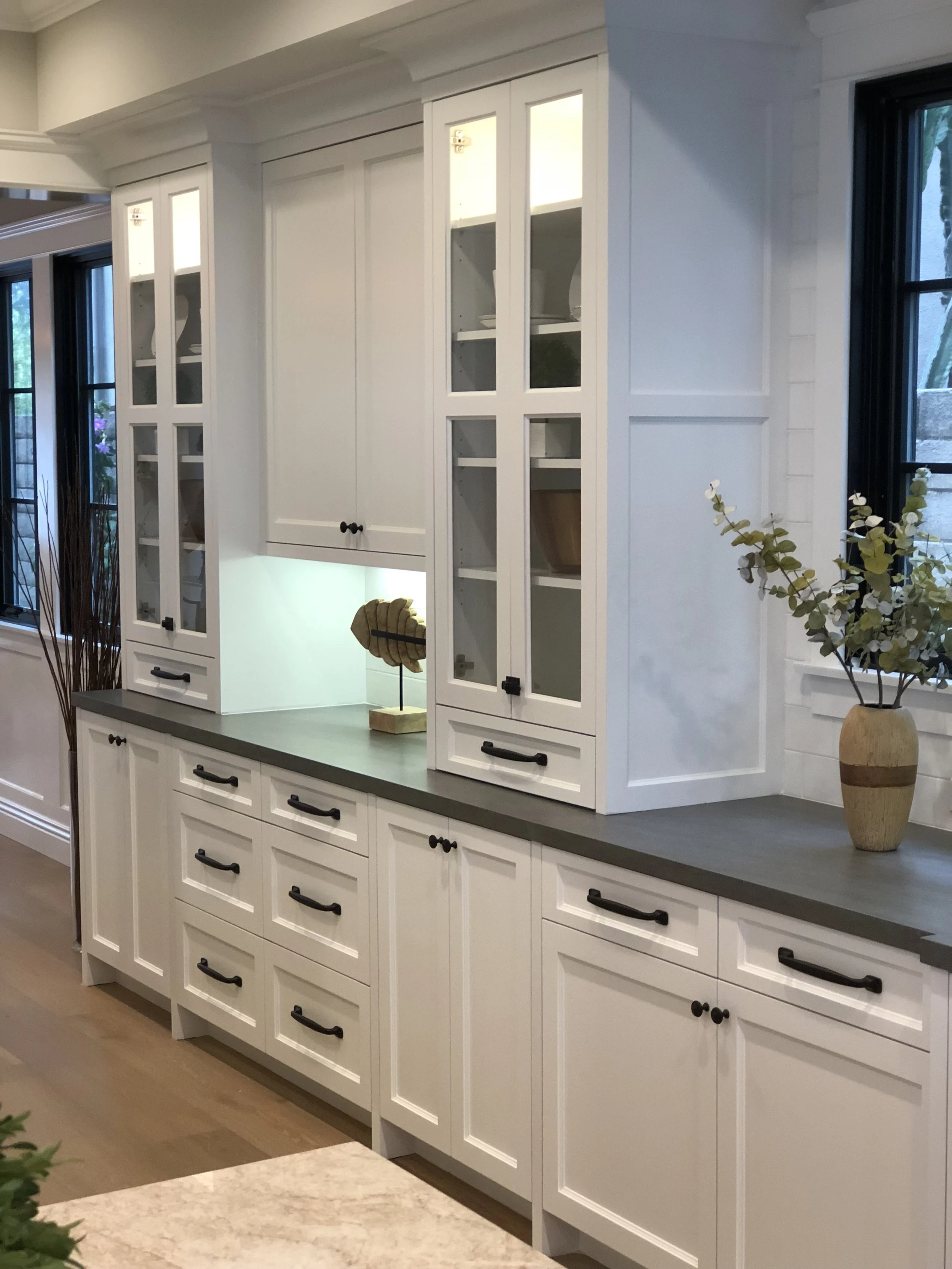 White kitchen cabinets with black handles, a gray countertop, a display shelf with decorative items, and a window with a vase of eucalyptus.