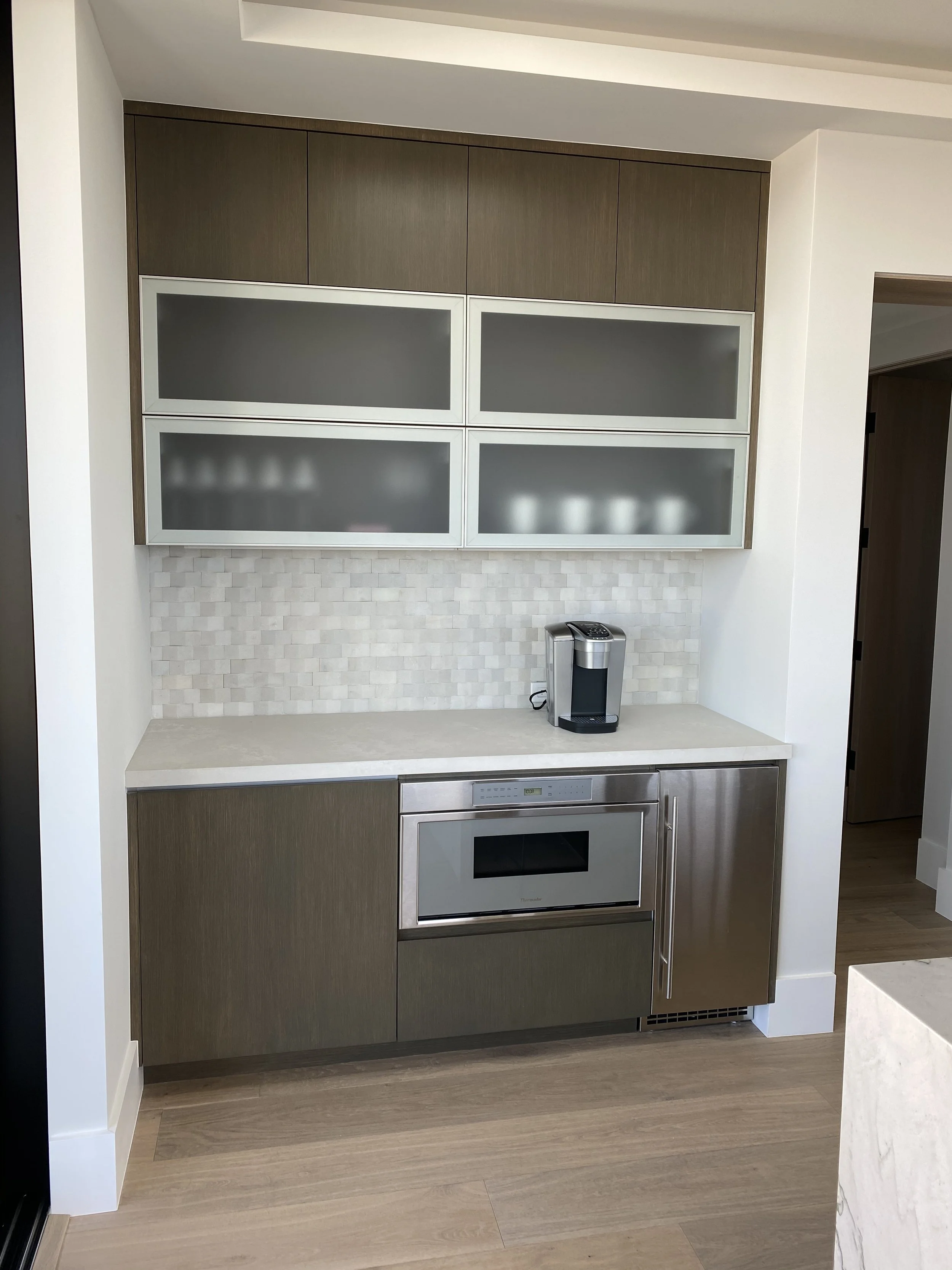 Modern kitchen with upper and lower cabinets, a built-in microwave, beige countertop, backsplash, and a coffee maker.