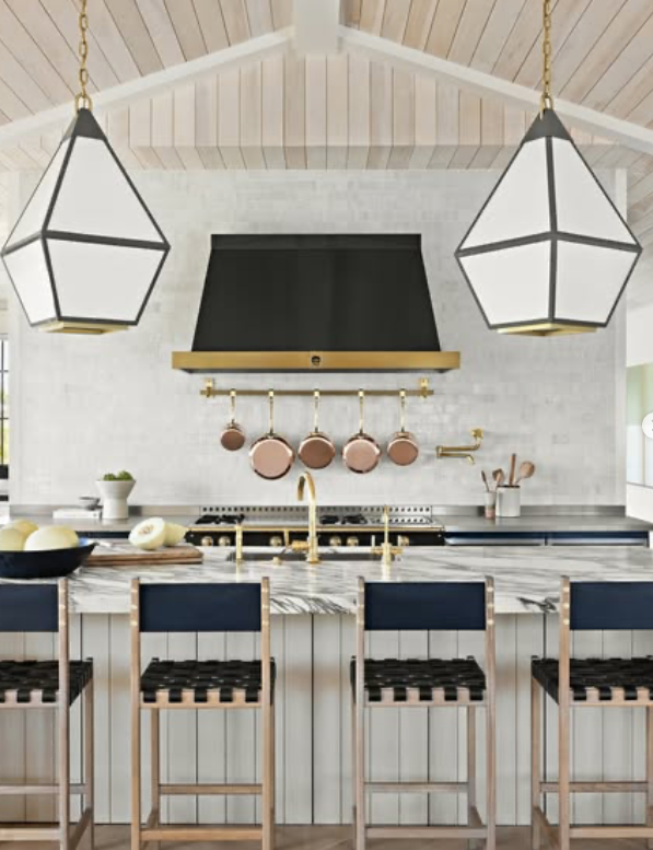Modern kitchen with white cabinetry, a marble island, copper pots hanging on a gold rail above the stove, black and white chairs at the breakfast bar, and two geometric pendant lights.