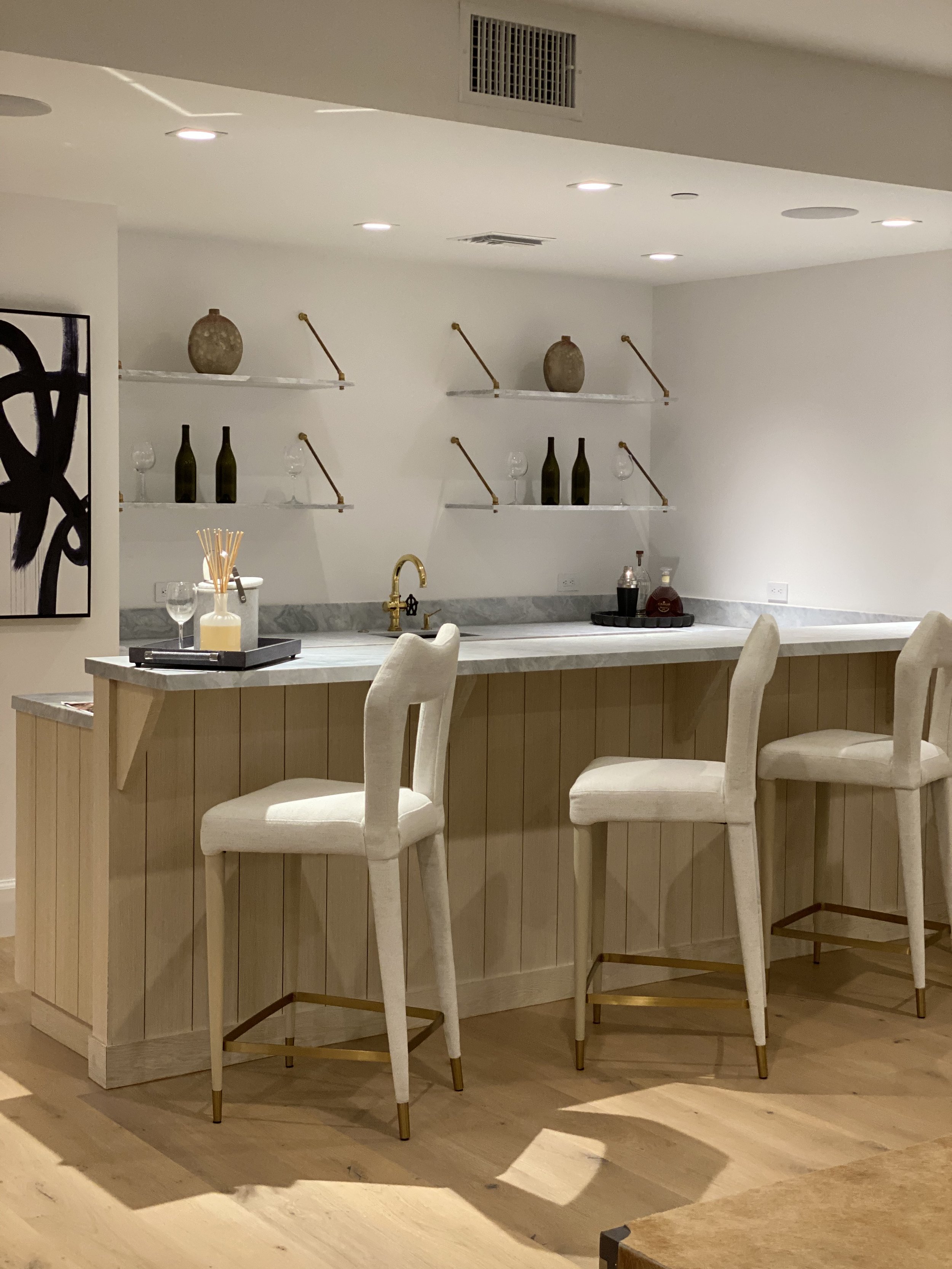 Modern home bar with beige upholstered bar stools, marble countertop, gold faucet, and open shelves with decorative vases and bottles.