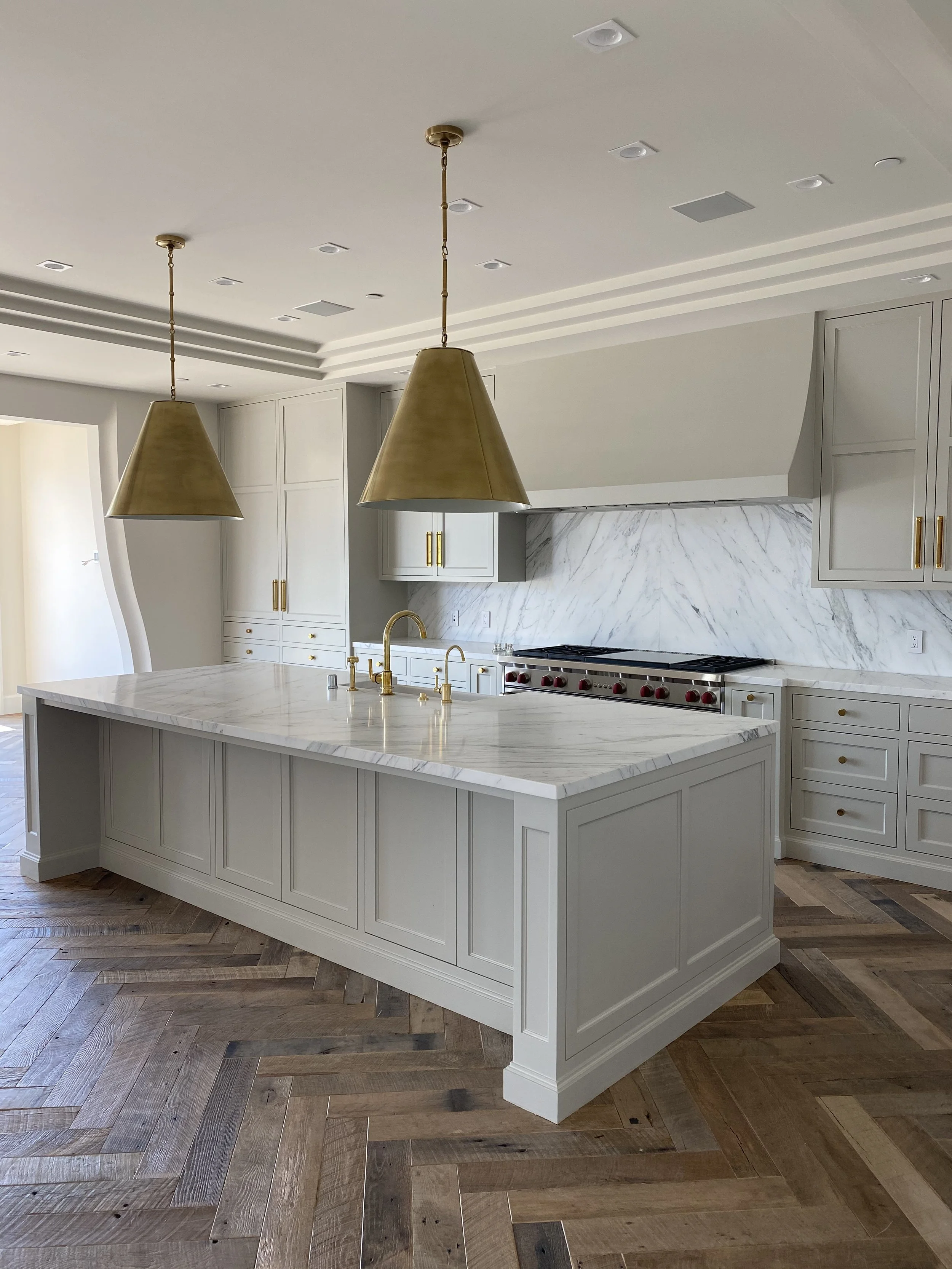 Modern kitchen with white cabinetry, marble backsplash, marble island, and gold fixtures. Brass pendant lights hang above the island, and there is a wooden herringbone floor.