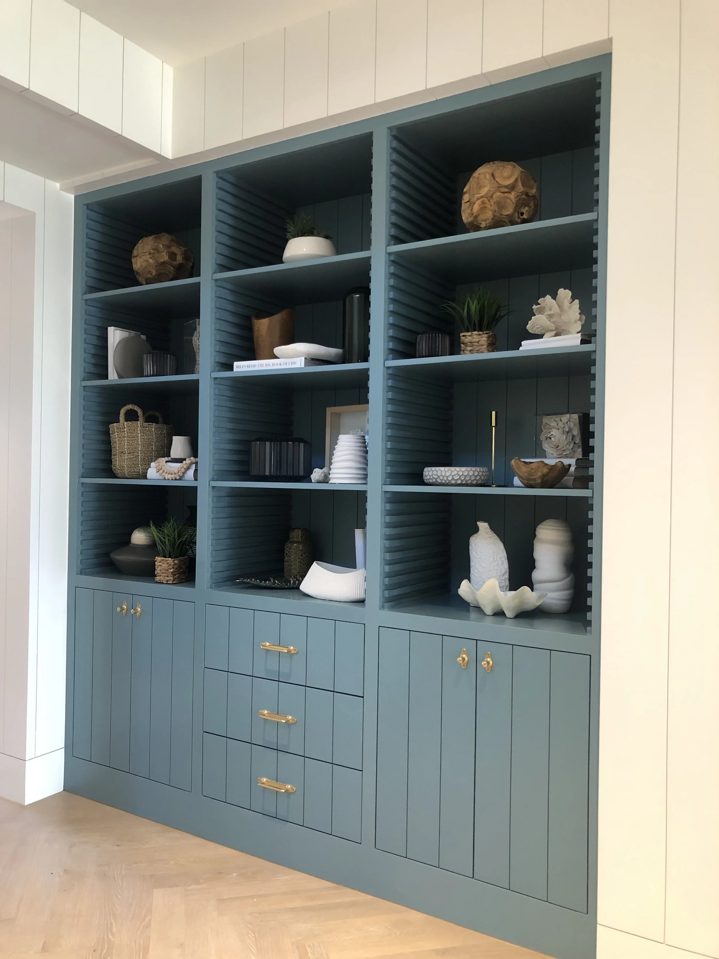 A blue built-in shelving unit with various decorative objects, including plants, vases, books, and sculptures, set against a white wall and above a light wood floor.